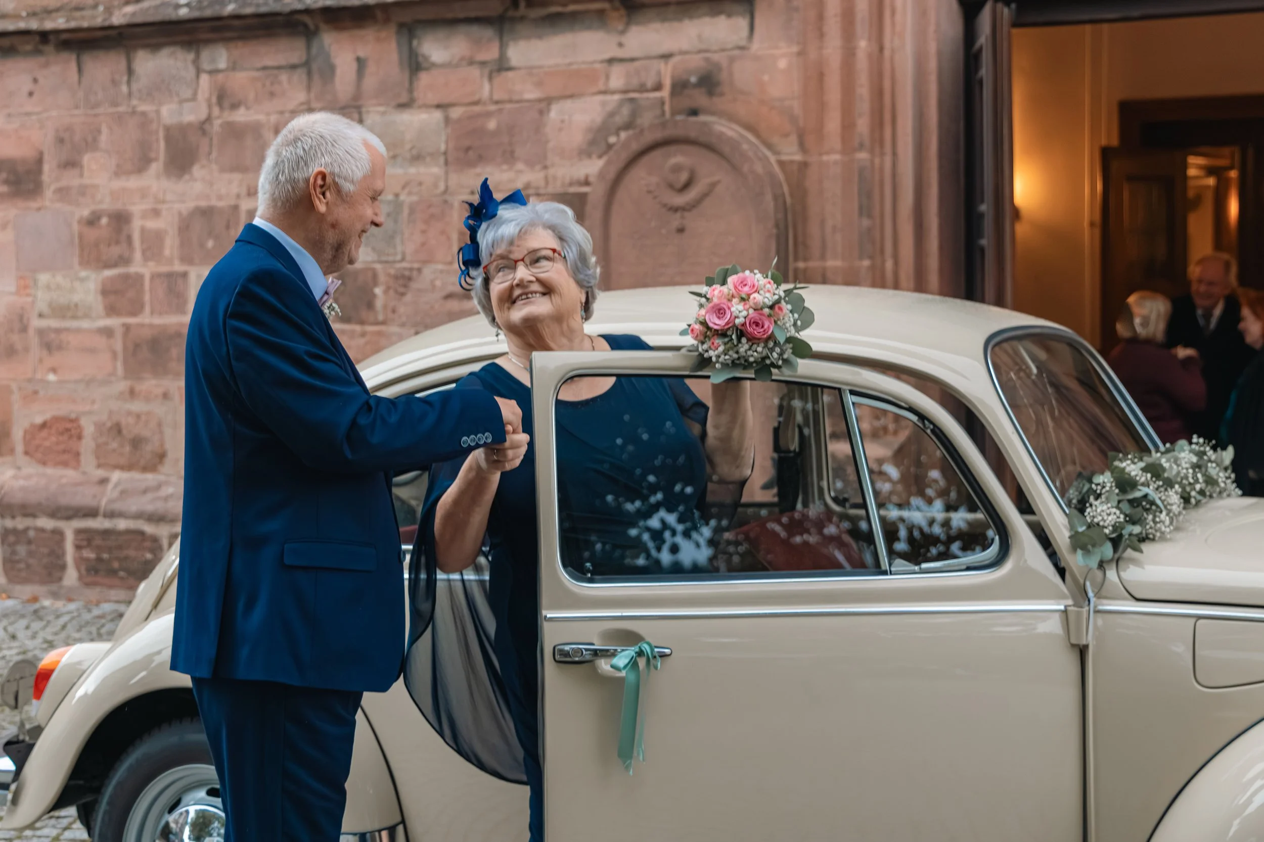 Ein älteres Paar, der Mann in einem blauen Anzug, die Frau in einem blauen Kleid mit einer blauen Schleife im Haar, stehen neben einem weißen Oldtimer mit Blumen und halten Händchen vor einer Kirche.