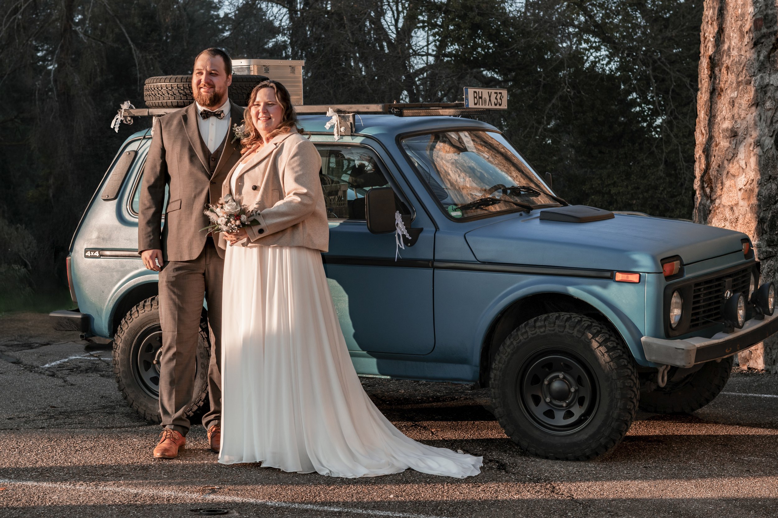 Ein Paar in formeller Kleidung steht vor einem blauen, alten Geländewagen, vermutlich bei Sonnenuntergang, bei einer Hochzeit oder besonderen Anlässen.