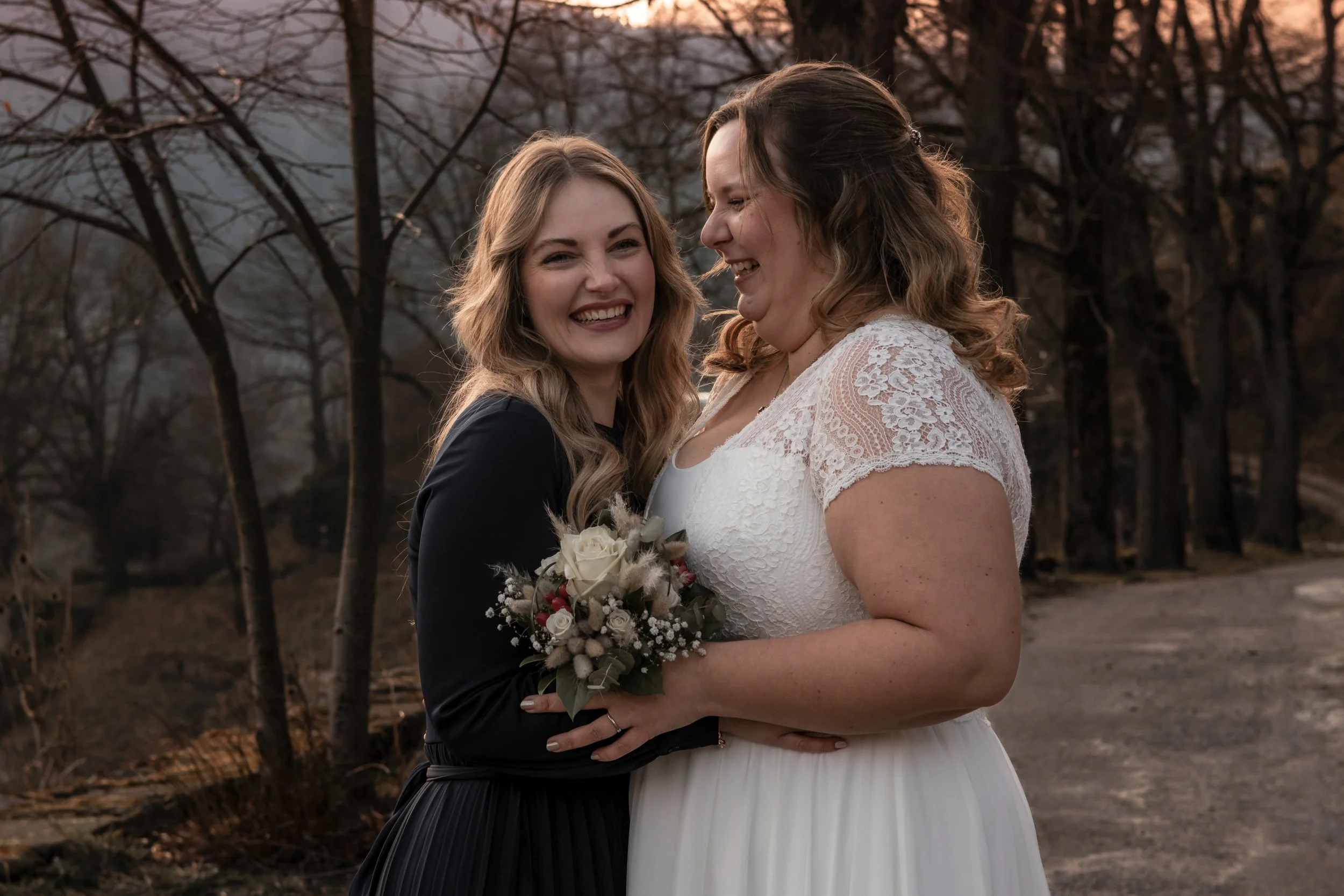 Zwei Frauen, eine in einem weißen Brautkleid und die andere in dunkler Kleidung, lachen und umarmen sich draußen bei Sonnenuntergang, eine hält einen Blumenstrauß.