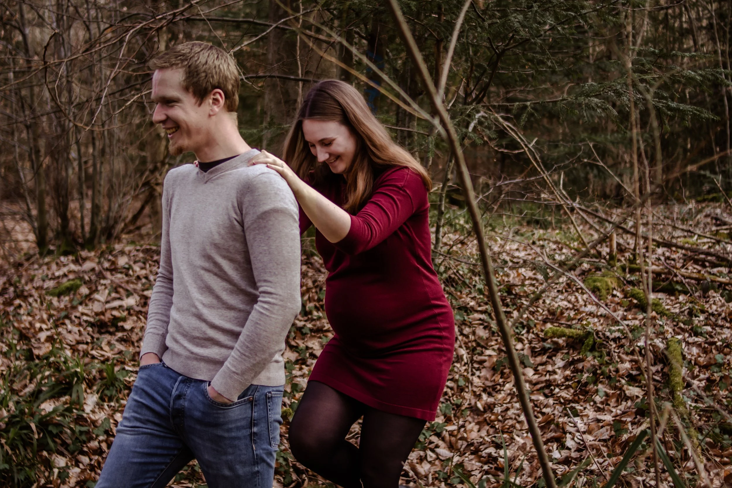 Ein lachendes Paar in einem Wald, er trägt graues Sweatshirt und Jeans, sie trägt ein rotes Kleid und schwarze Strumpfhosen, es ist Herbst.