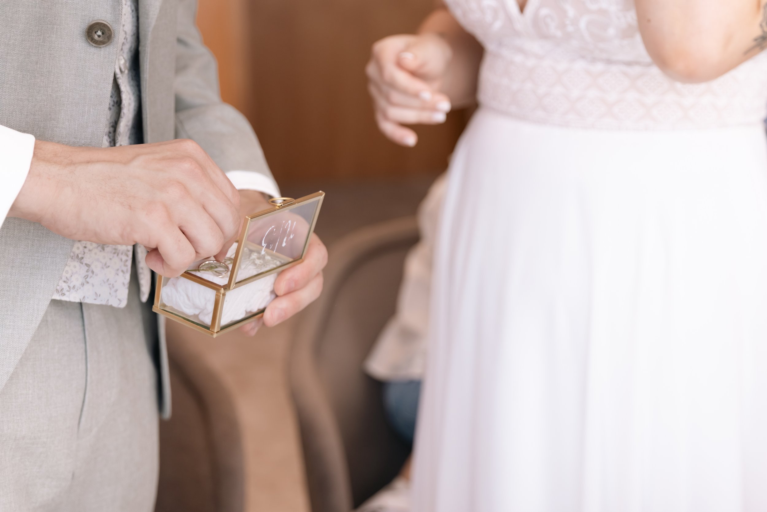 Ein Brautpaar bei der Trauung, das den Ehering austauscht. Der Bräutigam hält eine kleine Schachtel mit Ringen, während die Braut in einem weißen Kleid zu sehen ist.