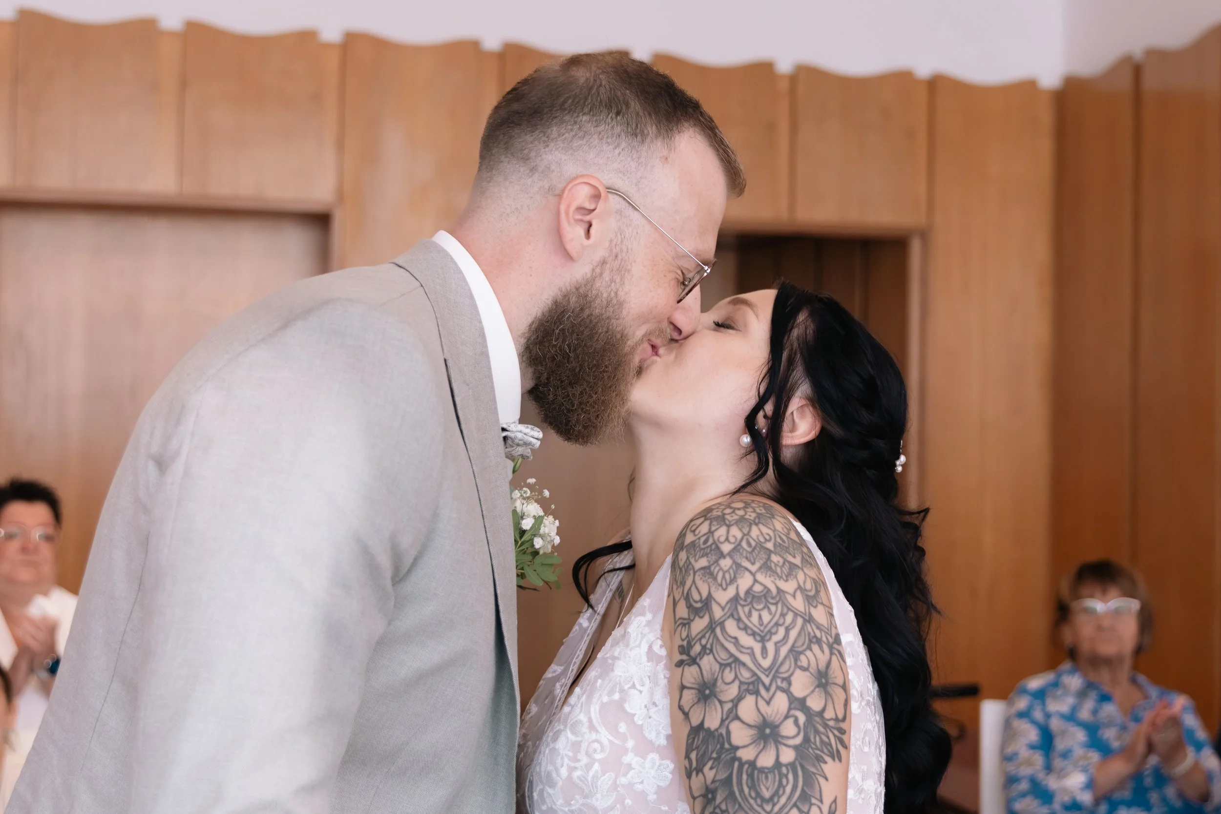 Ein Paar bei der Hochzeitszeremonie küsst sich. Der Mann trägt einen hellgrauen Anzug, die Frau hat schwarze Haare, Tattoo-Arm und trägt ein weißes Kleid.