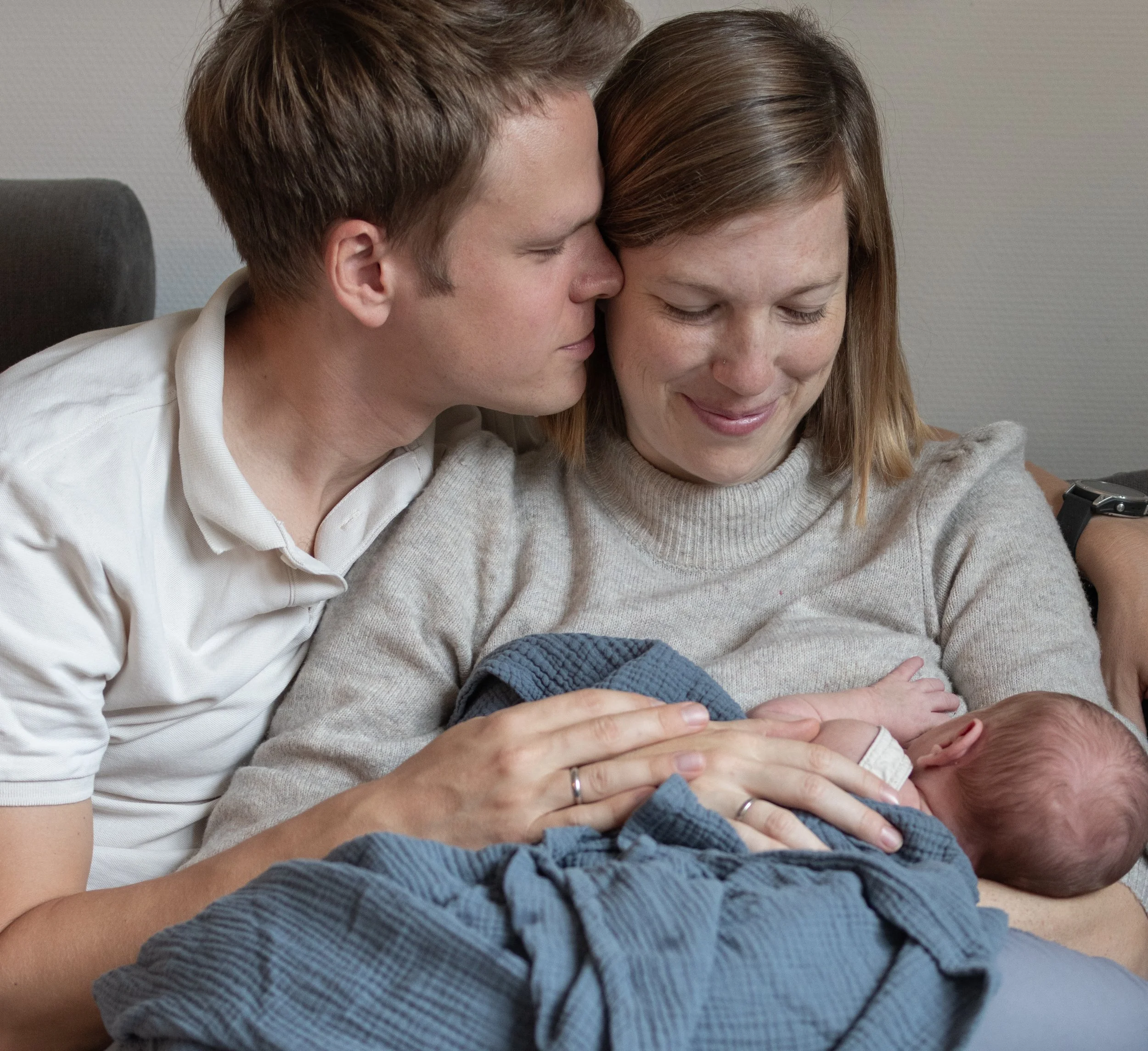 Eltern sitzen auf einer Couch, während die Mutter ein neugeborenes Baby stillt. Der Vater kuschelt sich an die Mutter und beide nehmen den Moment liebevoll wahr.