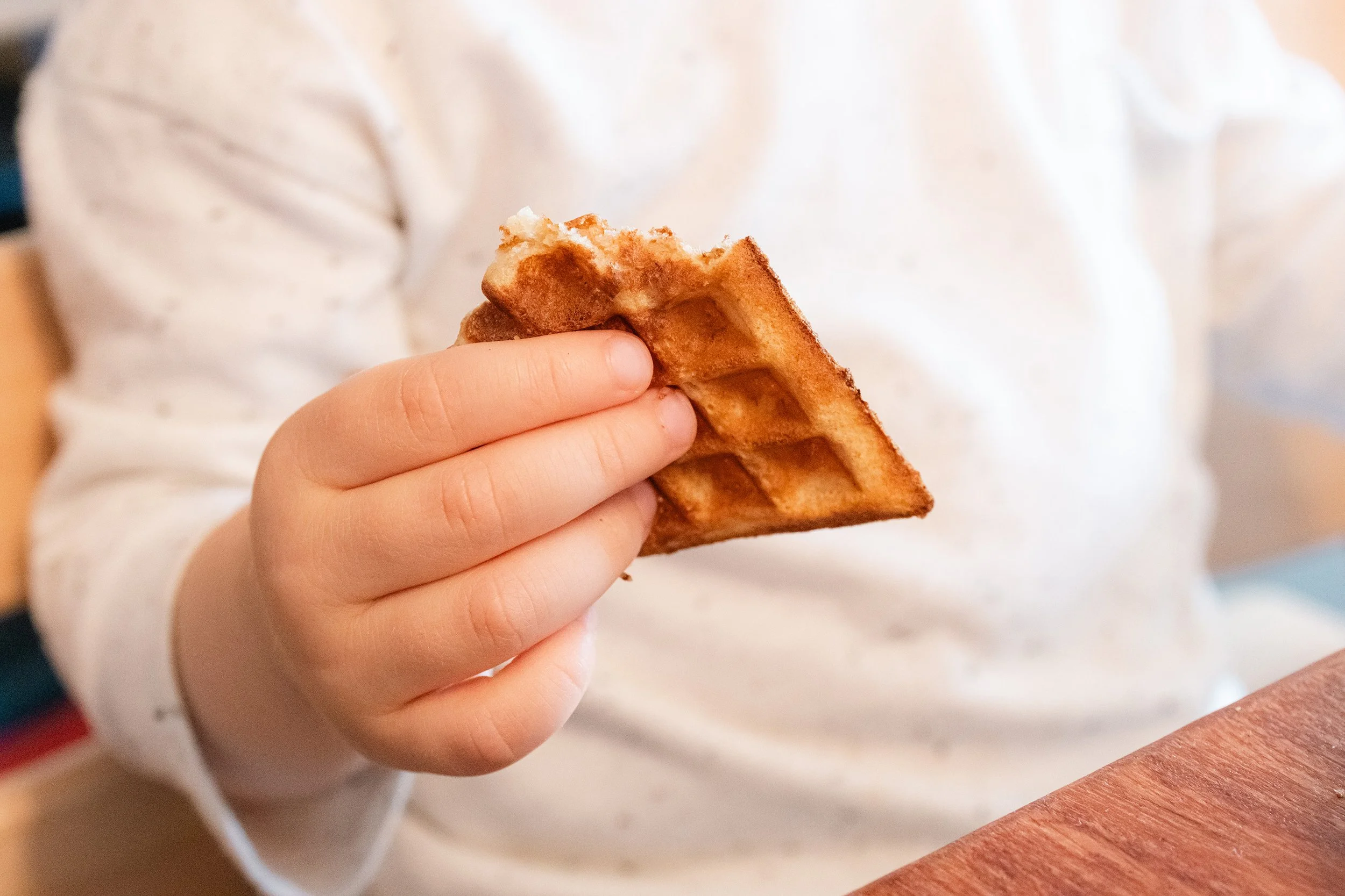 Nahaufnahme einer Hand, die eine goldbraune Waffel hält, während im Hintergrund eine Person in einem weißen Pullover sitzt.