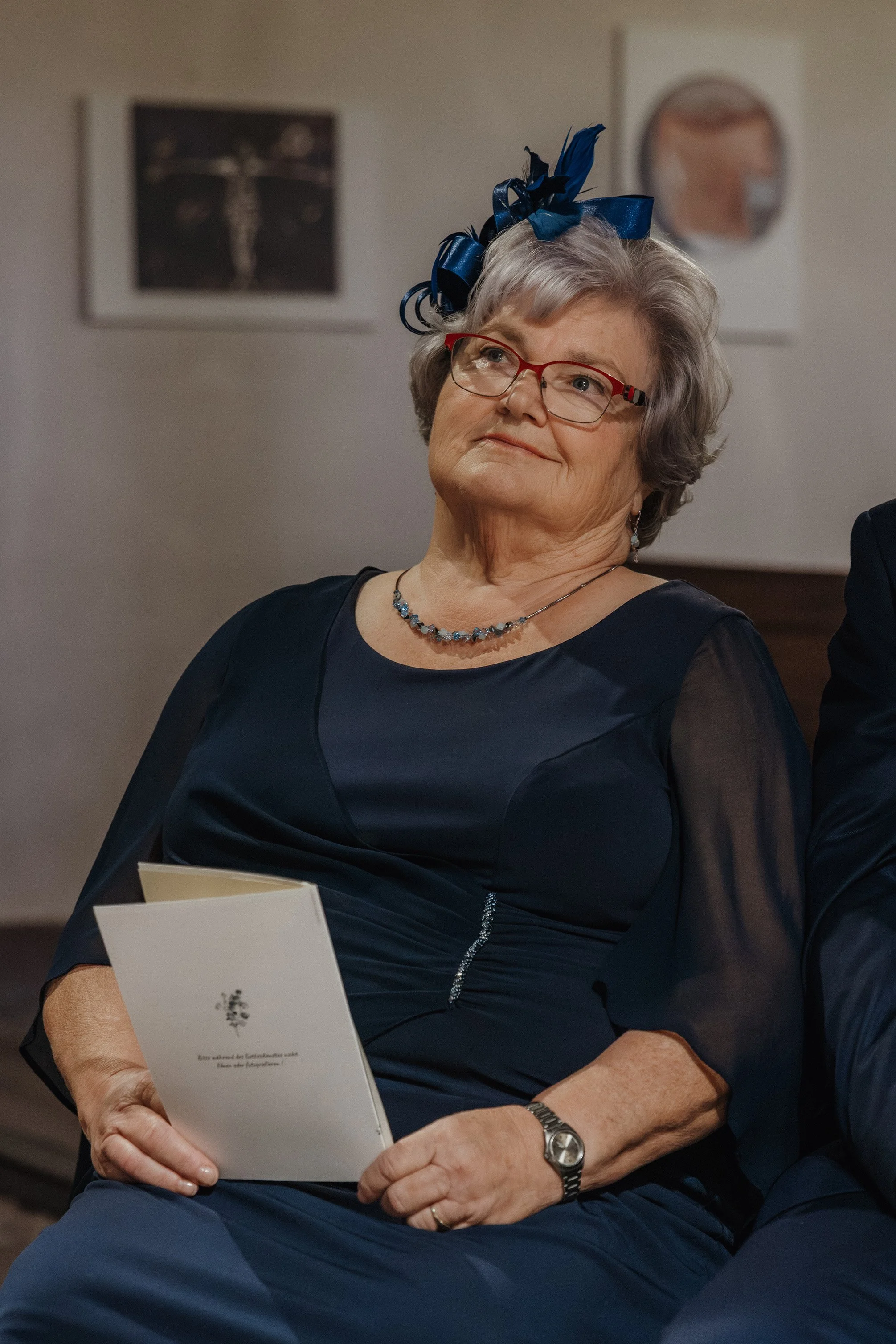 Ältere Frau in dunklem Kleid mit rotem Brillengestell, blauer Schleifen-Haarreif, Halskette, Armbanduhr und in der Hand eine Karte, vermutlich bei einer Feier, im Hintergrund Bilder an der Wand.