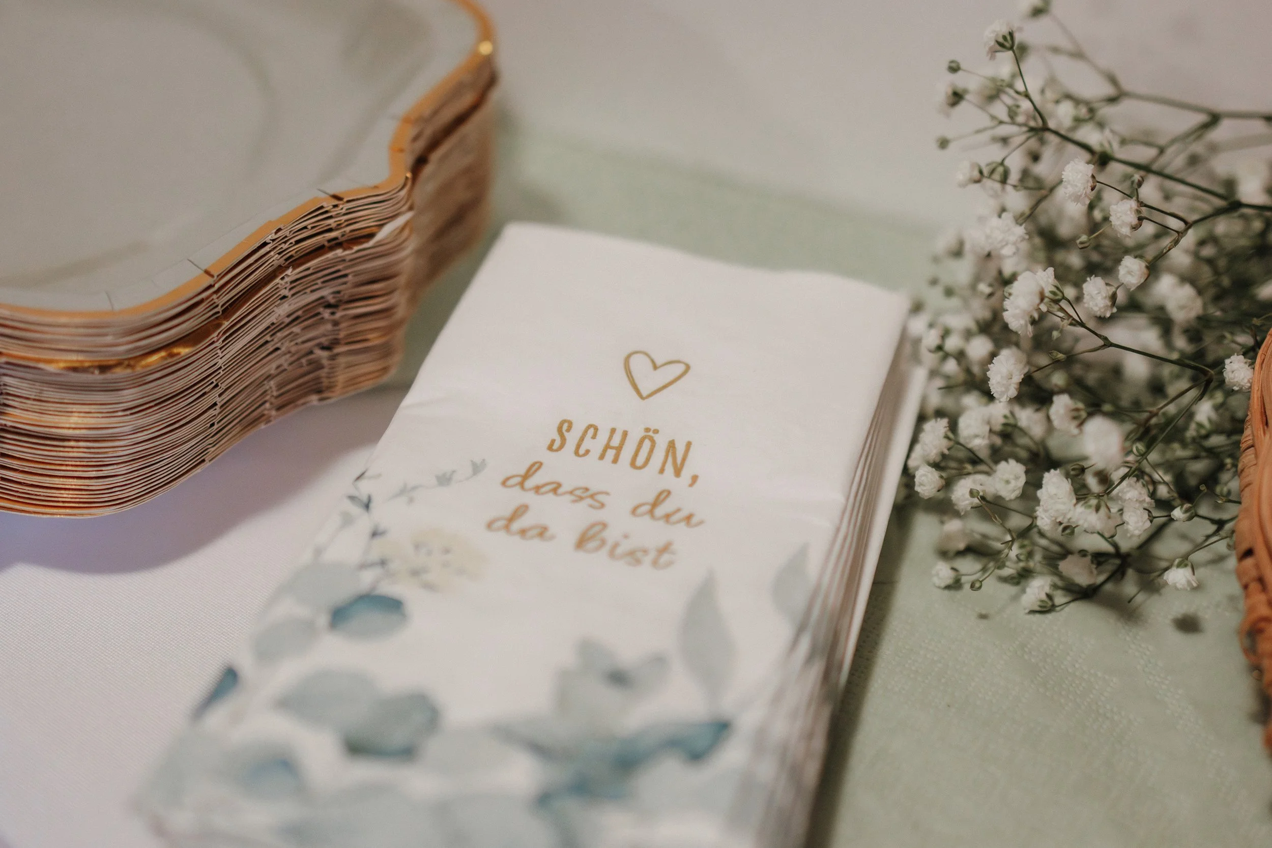 Serviette mit goldener Schrift und Herz, spruch: "Schön, dass du da bist" neben Lippenstift, Blumen und einem runden Korb.