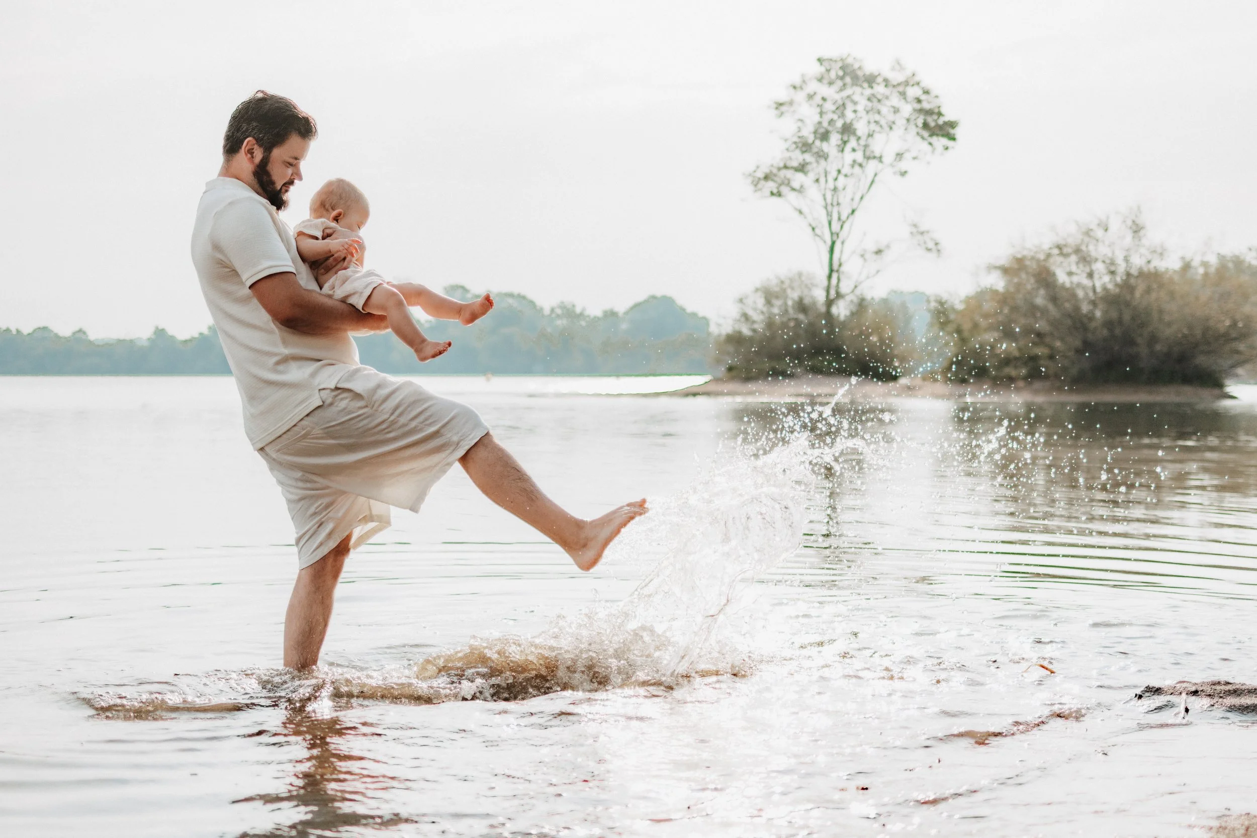 Vater und Kind spielen im Wasser an einem Flussufer, Vater hebt das Kind hoch während es im Wasser planscht.