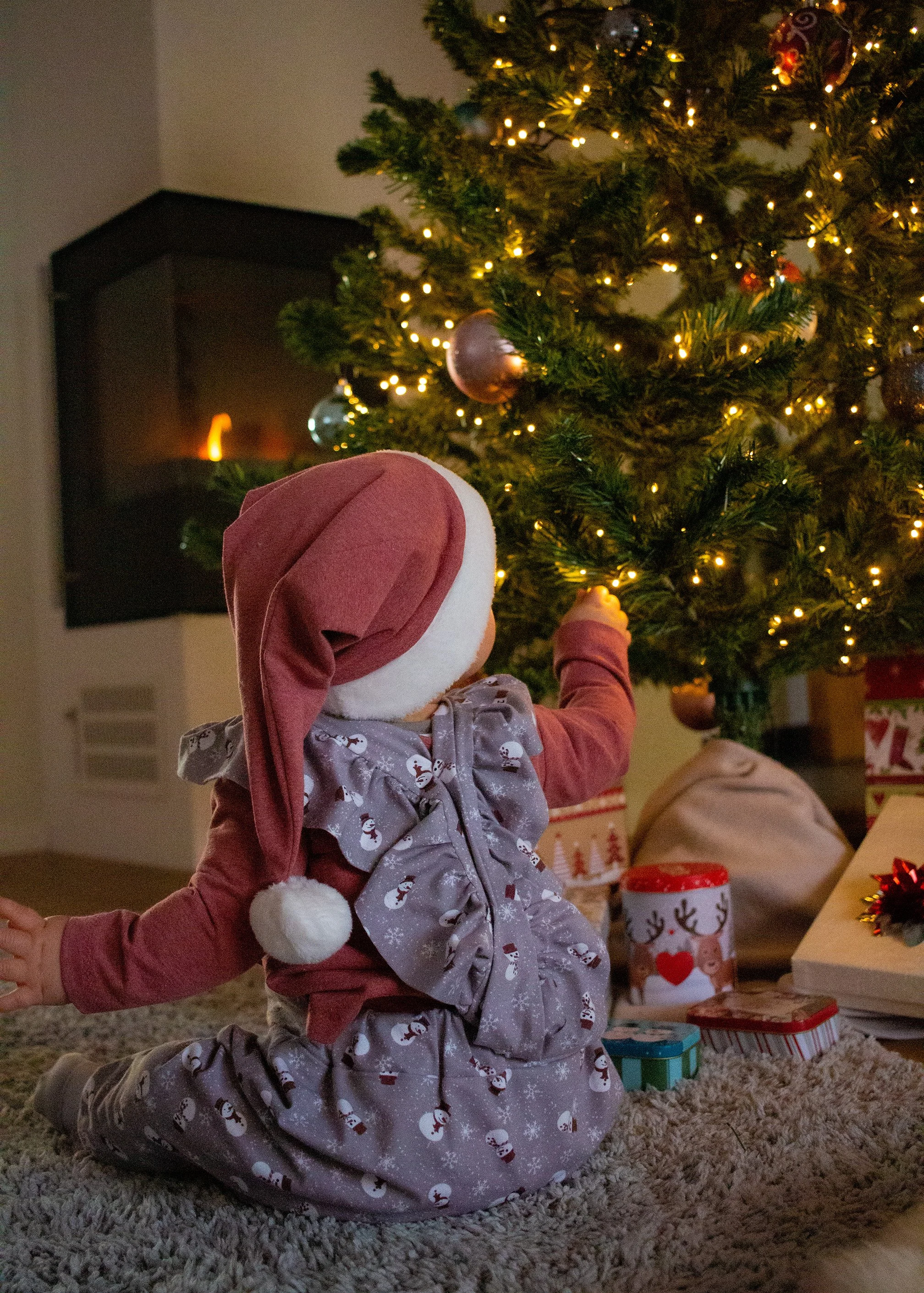 Ein Kind in Weihnachtskleidung sitzt vor einem geschmückten Weihnachtsbaum und berührt vorsichtig eine Lichterkette, umgeben von Geschenken.