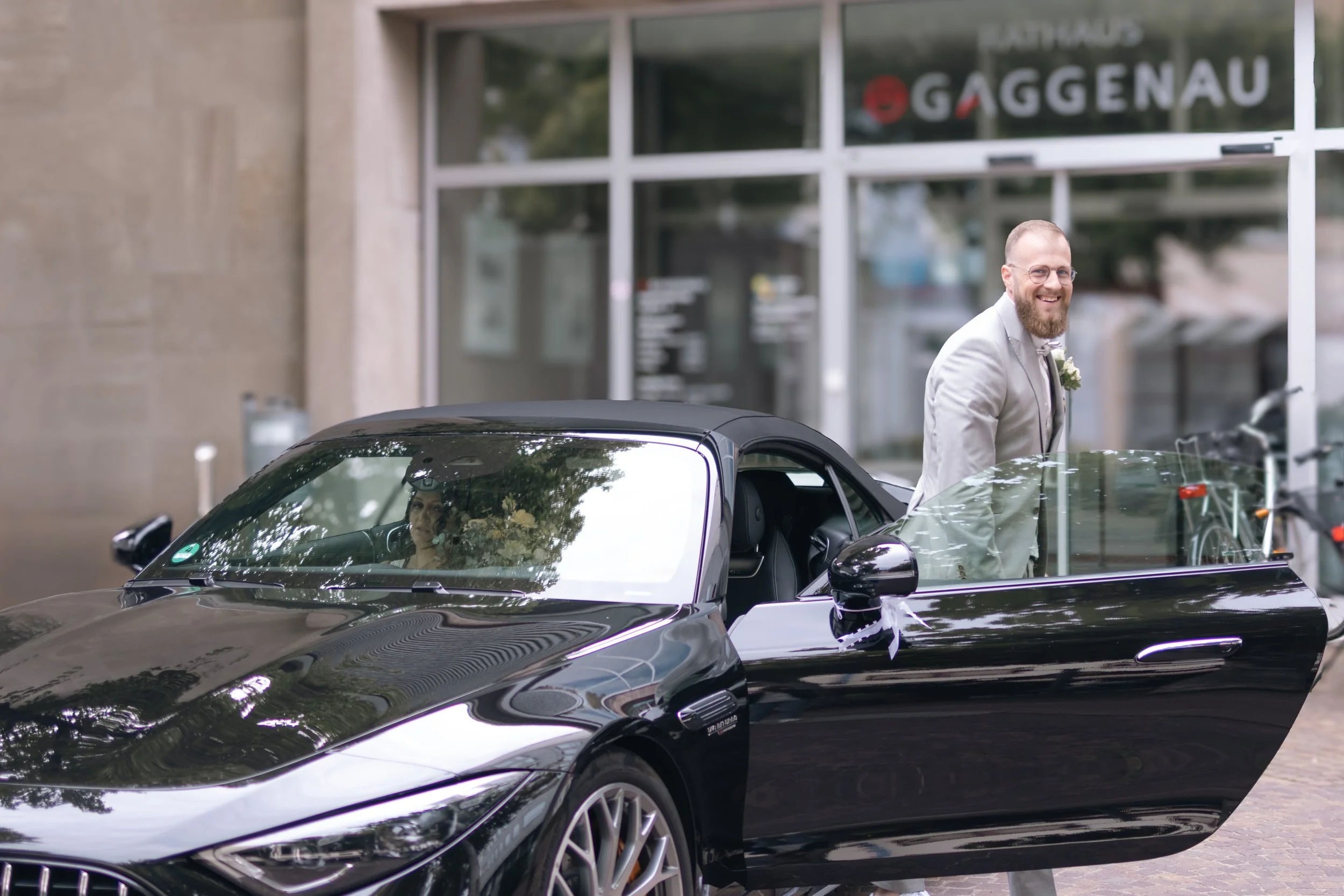 Ein Mann in einem hellen Anzug mit Fliege steht neben einem schwarzen Sportwagen, der vorne offen ist, vor einem Gebäude mit einem Schild "Gaggenau". Im Auto sitzt eine Frau, die nach vorne schaut, und die Außenspiegel sind mit weißen Bändern dekorie
