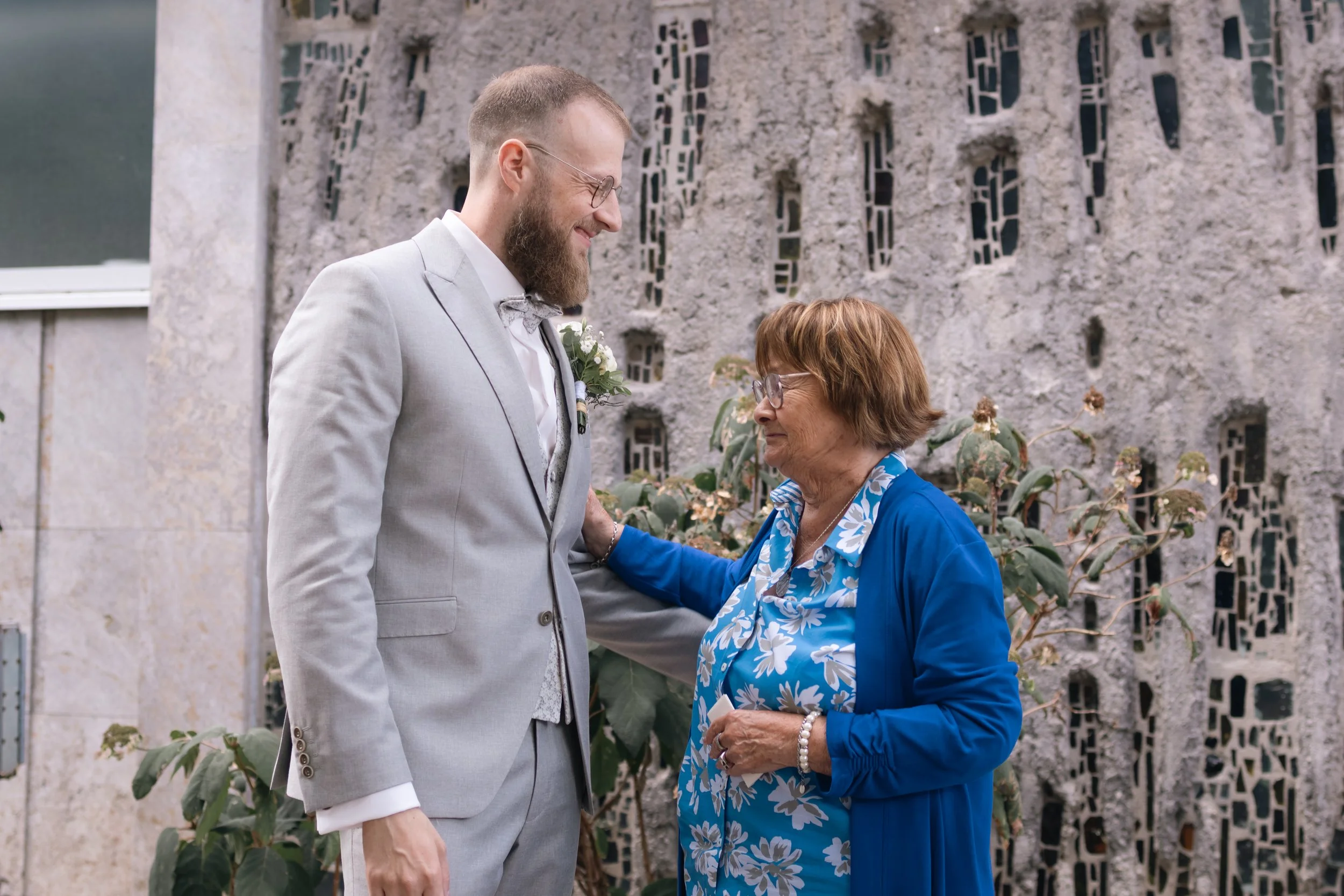 Ein junger Mann in einem hellgrauen Anzug, vermutlich bei einer Hochzeit, steht lächelnd mit einer älteren Frau, die eine blaue Bluse mit weißen Blumen und eine blaue Jacke trägt. Sie befinden sich vor einem modernen, künstlerisch gestalteten Betonma