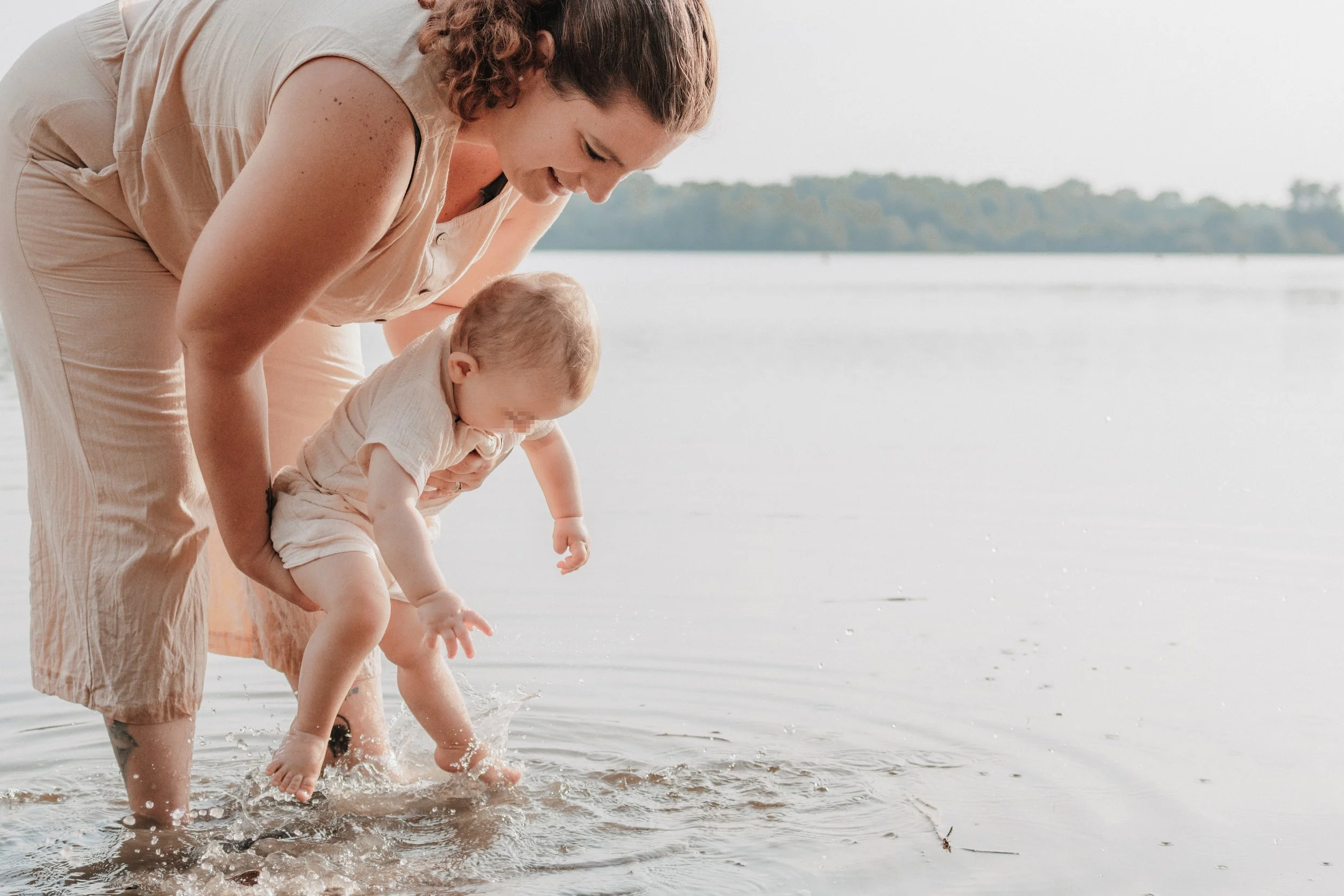 Eine Frau hält ein kleines Kind beim Spielen am Wasser eines Sees, während das Kind ins Wasser greift. Im Hintergrund sind Bäume und ein klarer Himmel sichtbar.