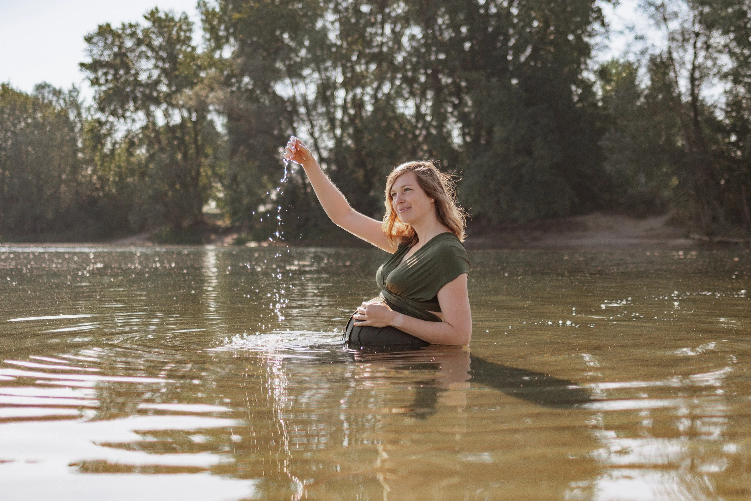 Junge Frau steht im Fluss und spielt mit Wasser, umgeben von Bäumen, bei Sonnenuntergang.