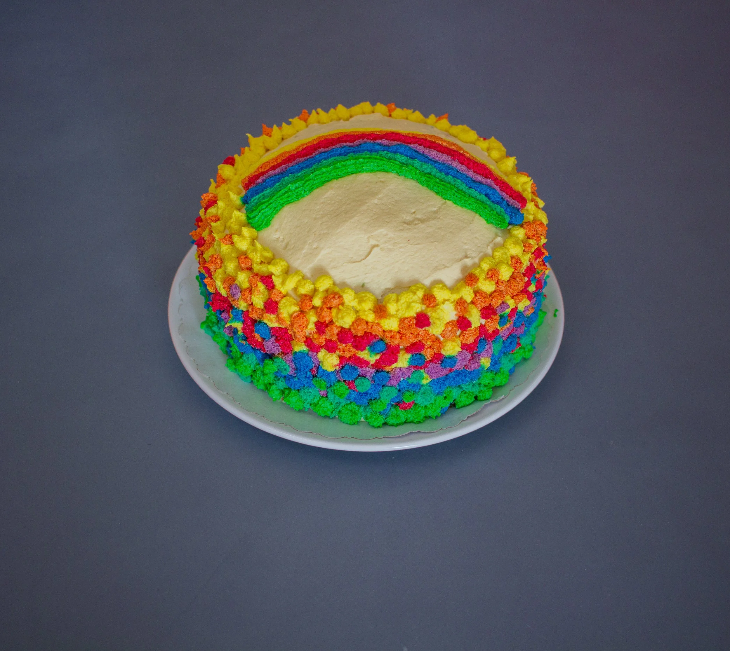 Ein runder Kuchen mit bunten Zuckerstreuseln in Regenbogenfarben und einem Bereich ohne Belag, auf einem weißen Teller vor einem grauen Hintergrund.