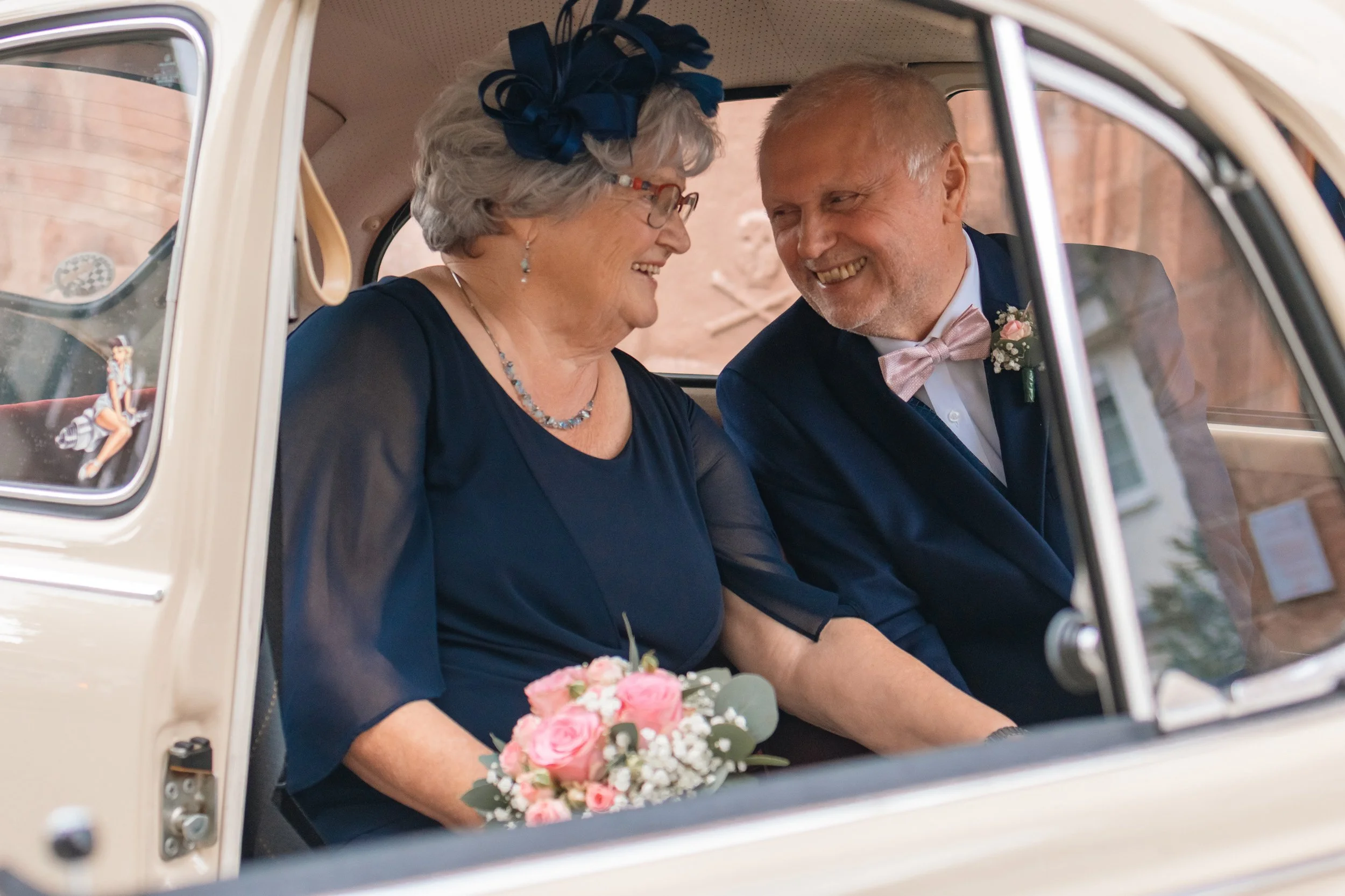 Eine ältere Frau und ein Mann sitzen in einem weißen Oldtimer, lachen und schauen sich liebevoll an. Die Frau hält einen kleinen Blumenstrauß in den Händen.