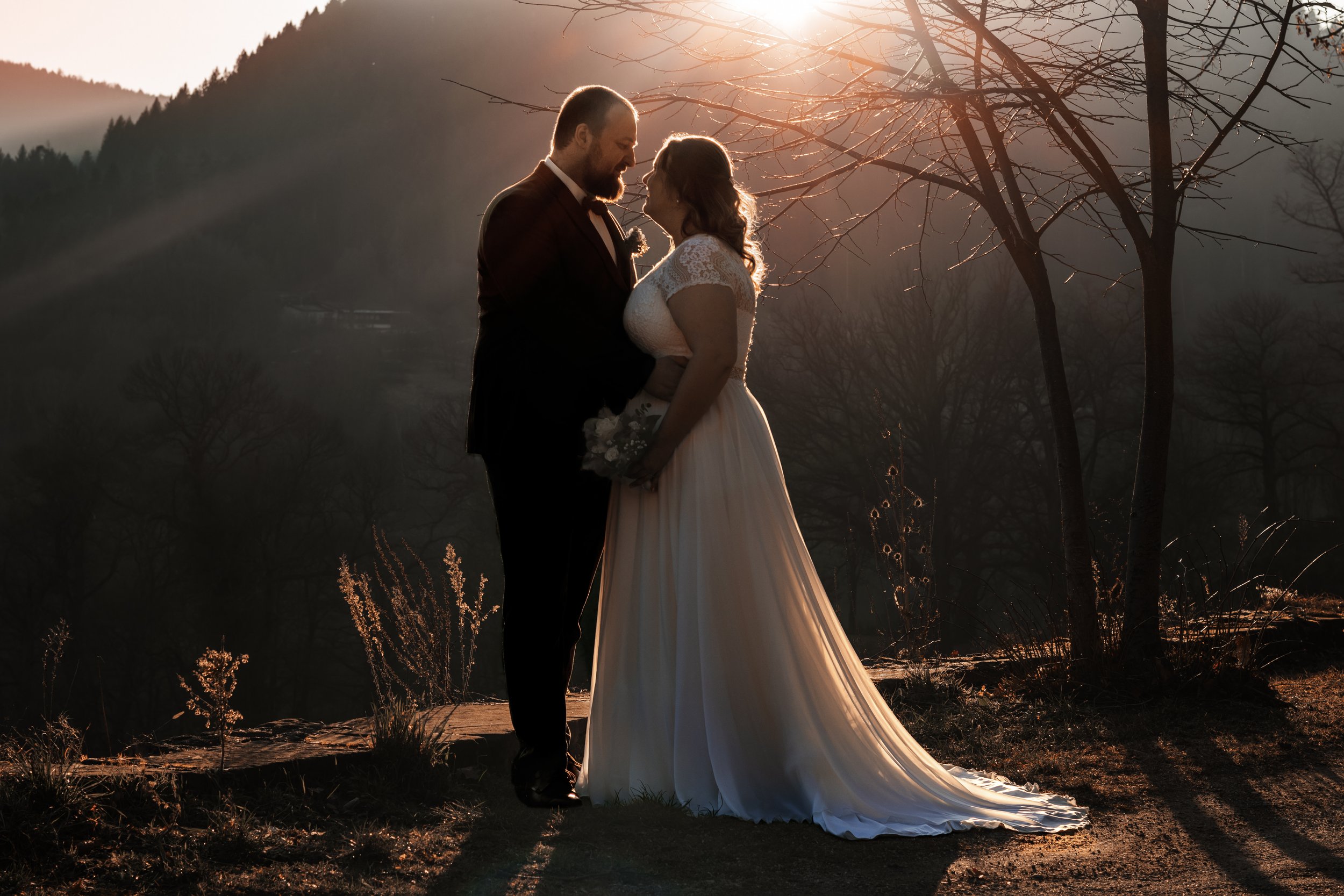 Ein Brautpaar steht sich bei Sonnenuntergang gegenüber, hält sich an den Händen und zeigt Liebe. Im Hintergrund sind Bäume und Berge sichtbar.