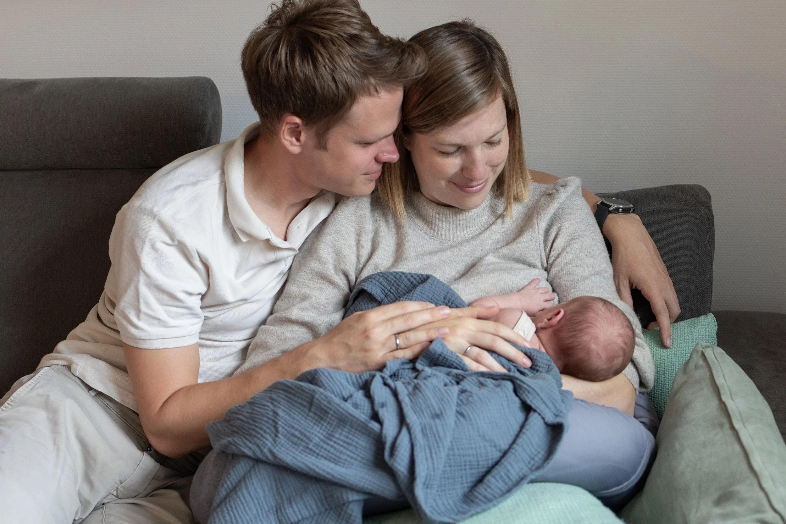 Ein Mann und eine Frau küssen ein Baby auf dem Sofa, während sie es liebevoll halten.
