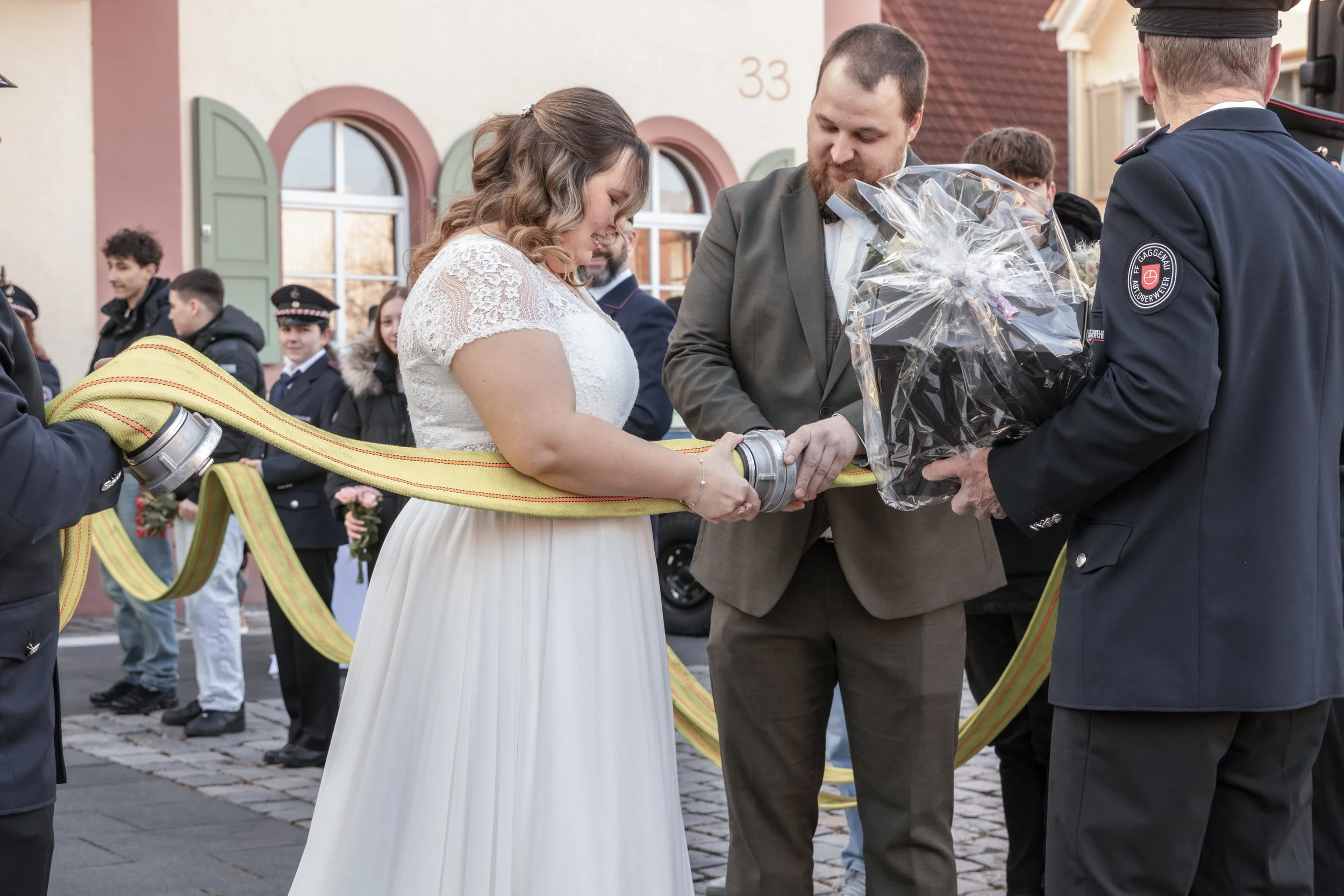 Braut und Bräutigam bei einer Feuerwehrhochzeit, die sich die Hände schütteln, während Feuerwehrleute im Hintergrund stehen, einige mit Blumen, vor einem Haus mit roten Ziegeln und grünen Fensterläden