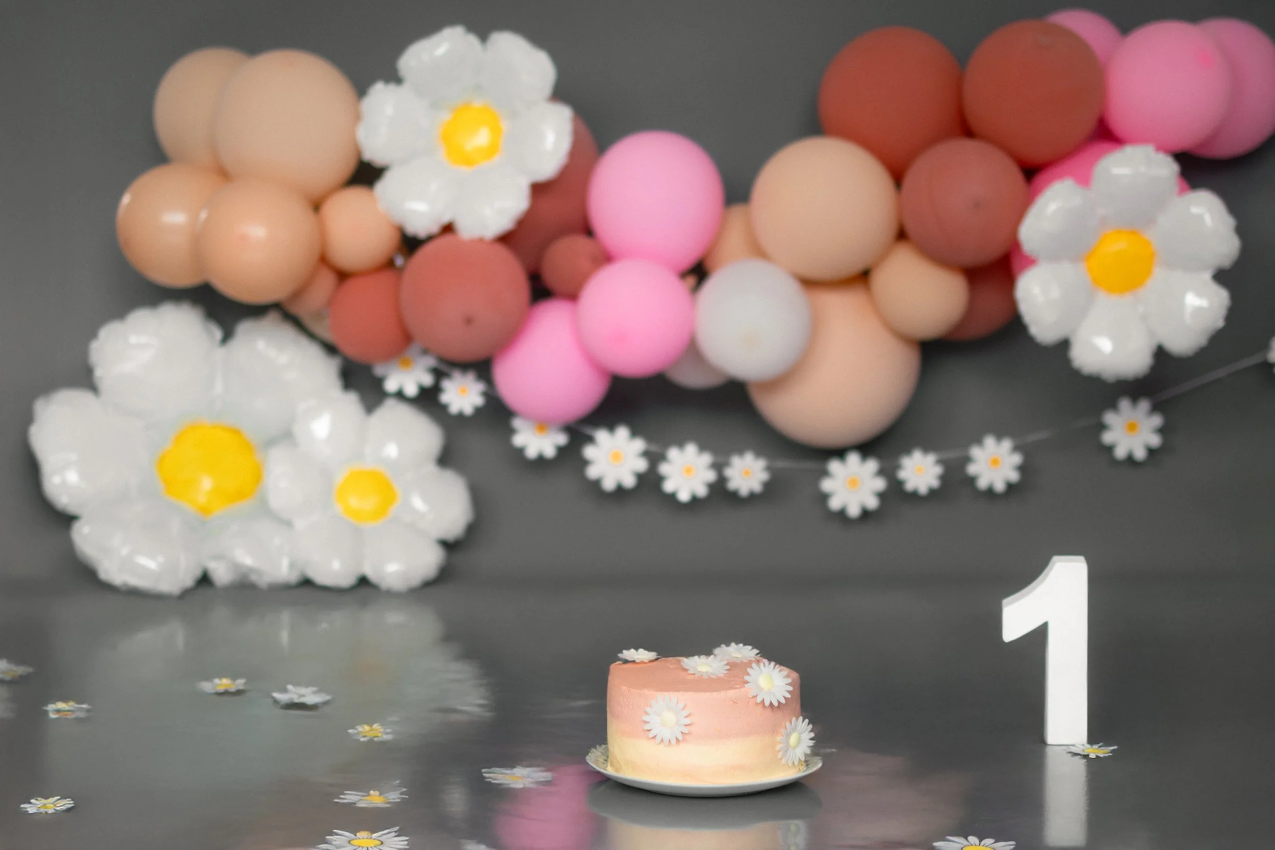 Kleine Torte mit Kamillendesign, im Vordergrund auf dem Tisch, daneben eine weiße Zahl 1. Im Hintergrund Bälle in Rosa, Beige und Rostrot, sowie Girlanden mit Kamillendesign, Dekoration für einen Kindertag.