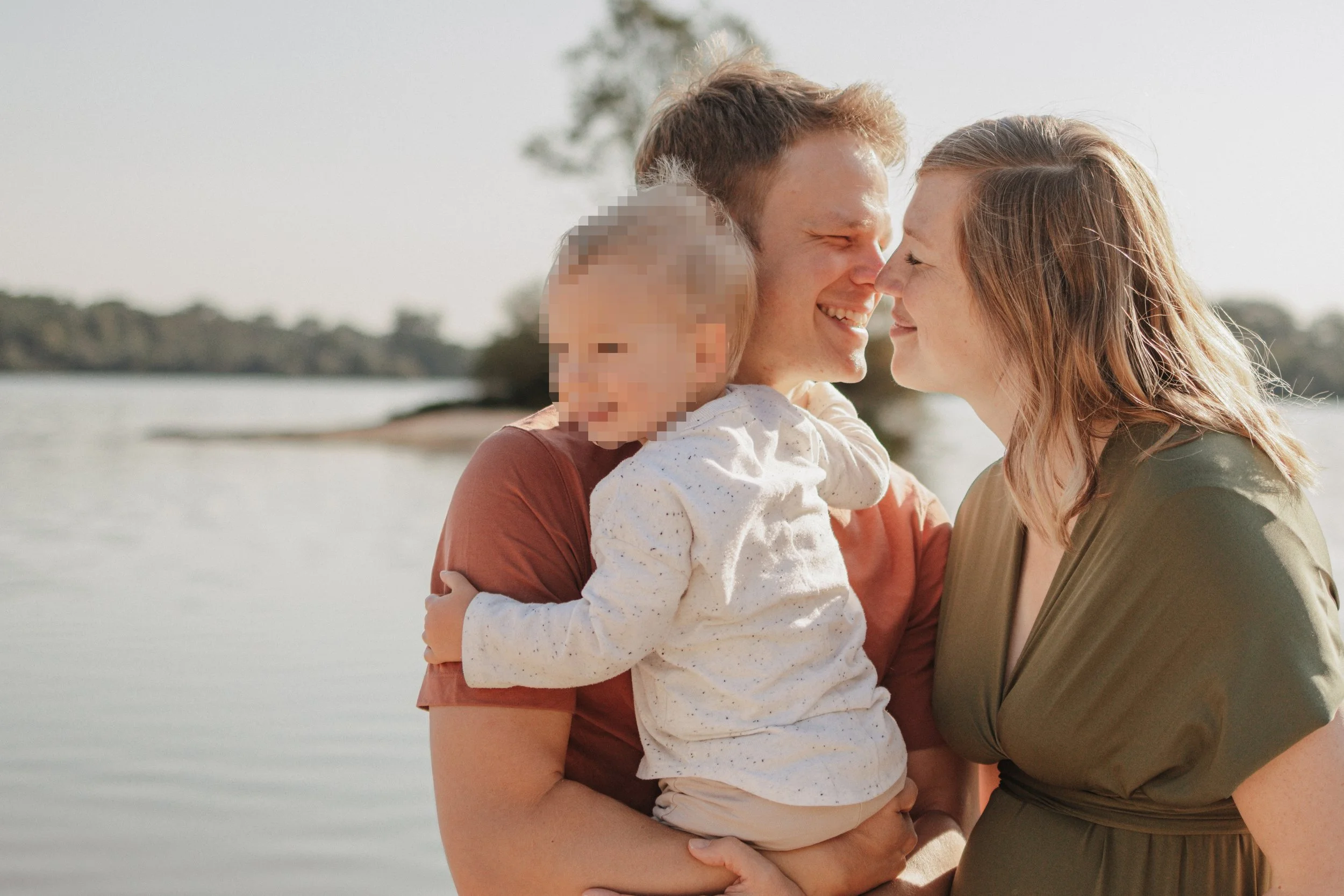Ein glückliches Familienfoto von einem Mann, einer Frau und einem kleinen Kind, die sich am Wasser in der Natur umarmen und lächeln.