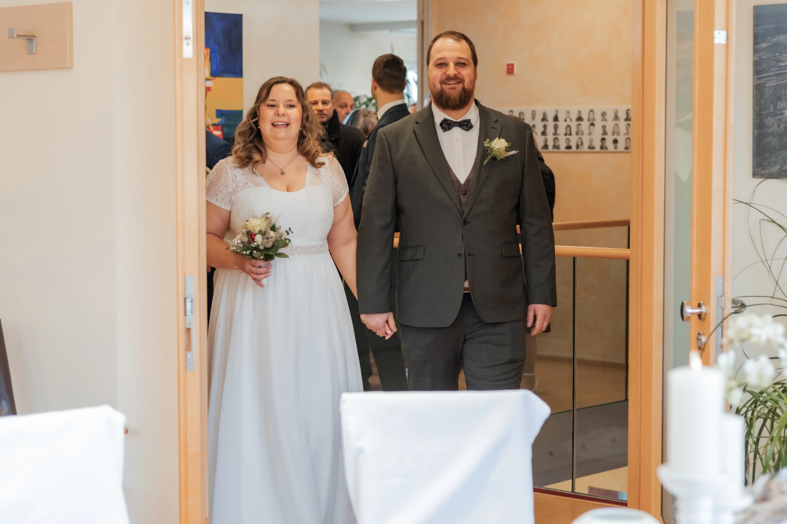 Ein Brautpaar, ein Mann in Anzug und eine Frau in Hochzeitstraum, hält die Hand des Partners und lächelt, während sie in einen Raum mit mehreren Gästen eintritt.