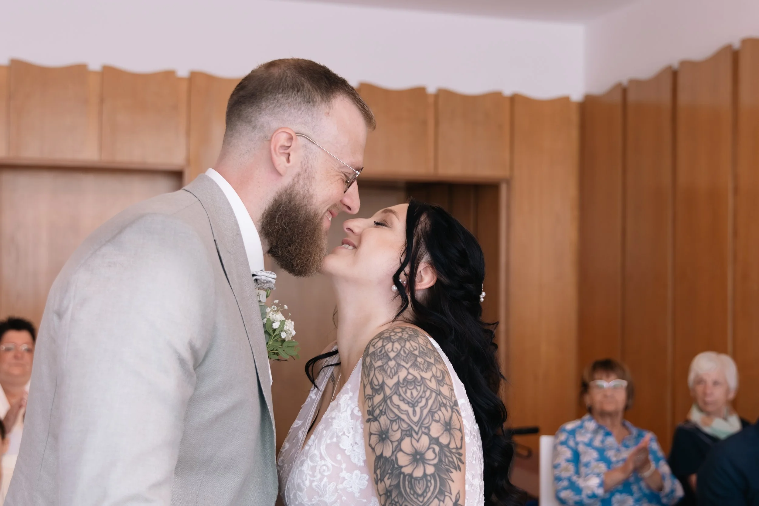 Ein frisch verheiratetes Paar steht sich bei ihrer Hochzeit gegenüber und berührt sich mit der Nase, während sie sich anlächeln, im Hintergrund sitzen Gäste in einem Saal.
