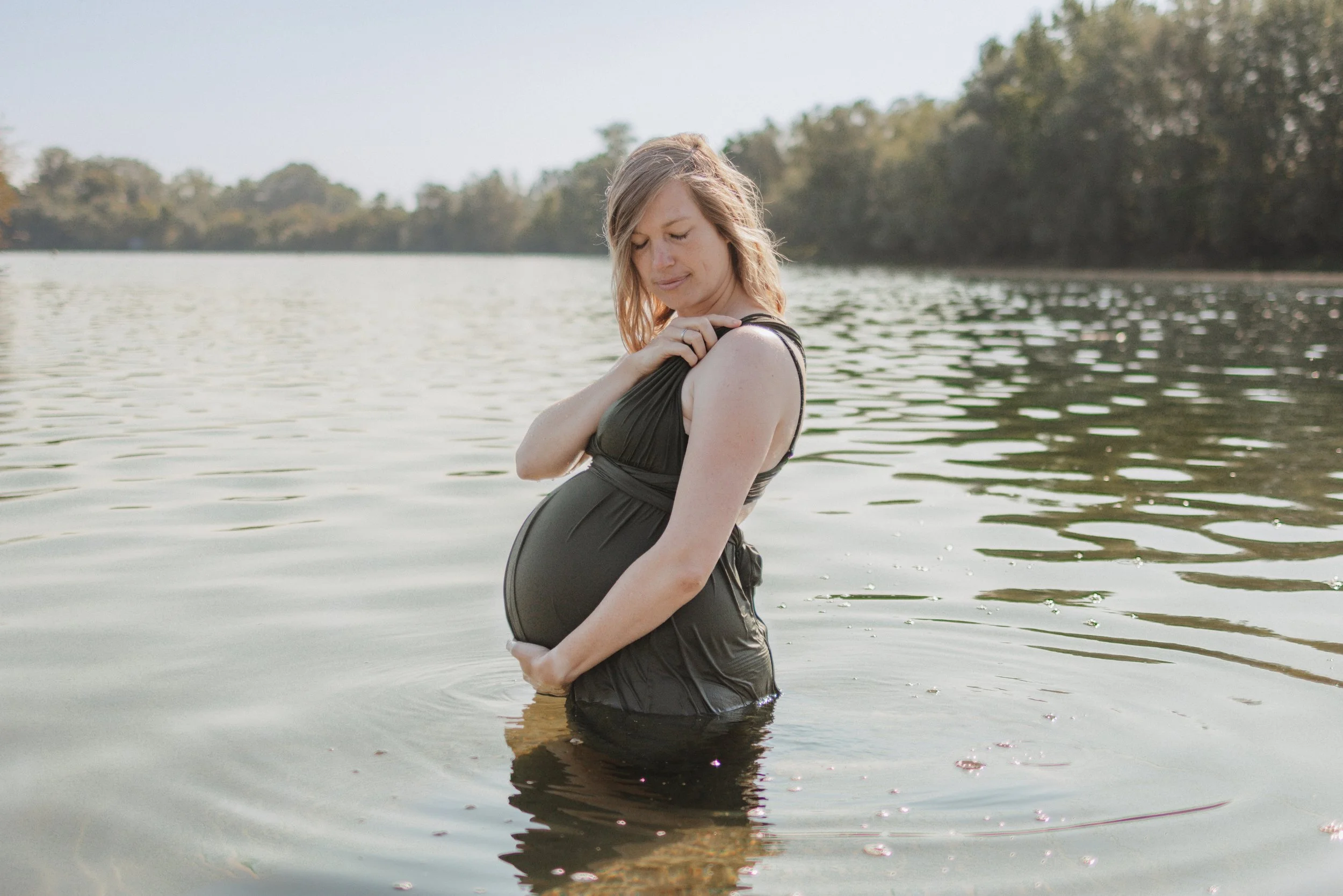Schwangere Frau steht im Wasser an einem Fluss oder See, hält ihren Bauch und schaut nachdenklich.