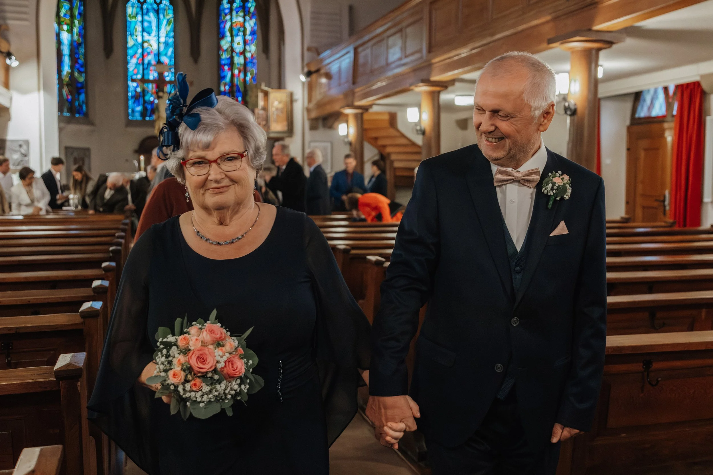 Älteres Paar bei einer Hochzeit in einer Kirche, Hand in Hand, lachend, Braut trägt eine dunkle Kleidung mit einem Blumenstrauß, der Bräutigam trägt einen Anzug mit Fliege, im Hintergrund weitere Gäste und bunte Buntglasfenster.