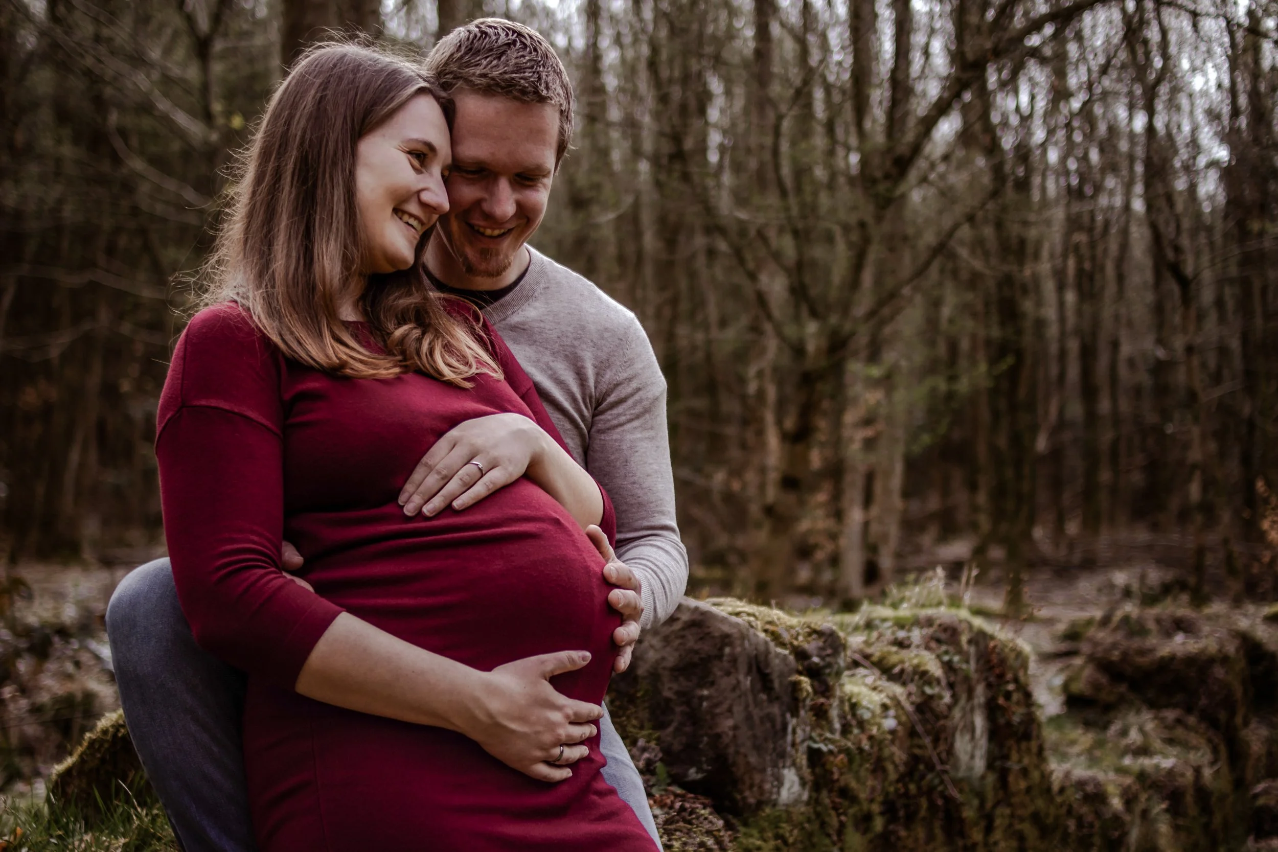 Ein Paar umarmt eine schwangere Frau im Wald, beide lachen und schauen sich liebevoll an.
