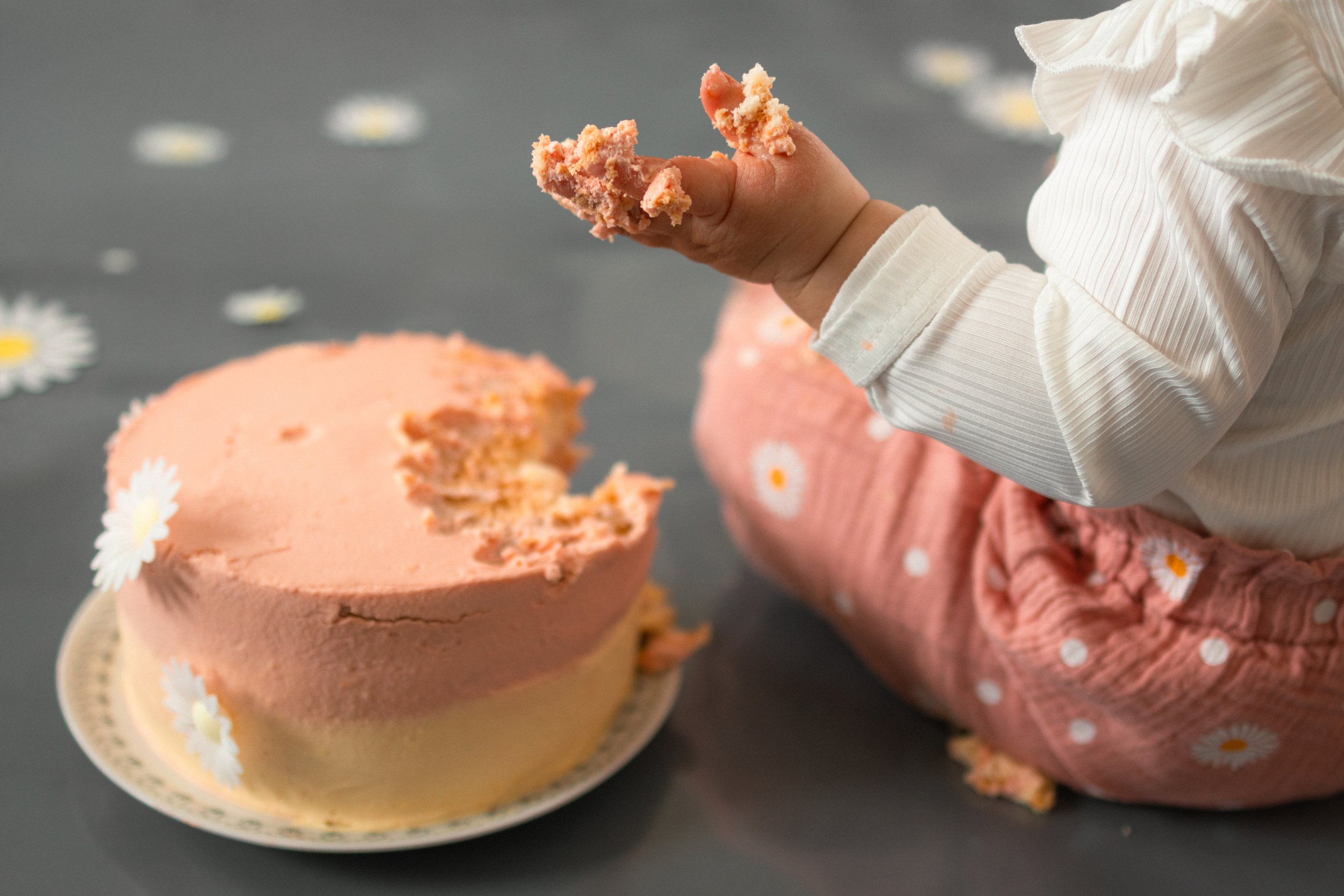 Ein Kind sitzt auf dem Boden und isst Kuchen. Der Kuchen ist halb gegessen, rosa und mit weißen Blumen dekoriert.