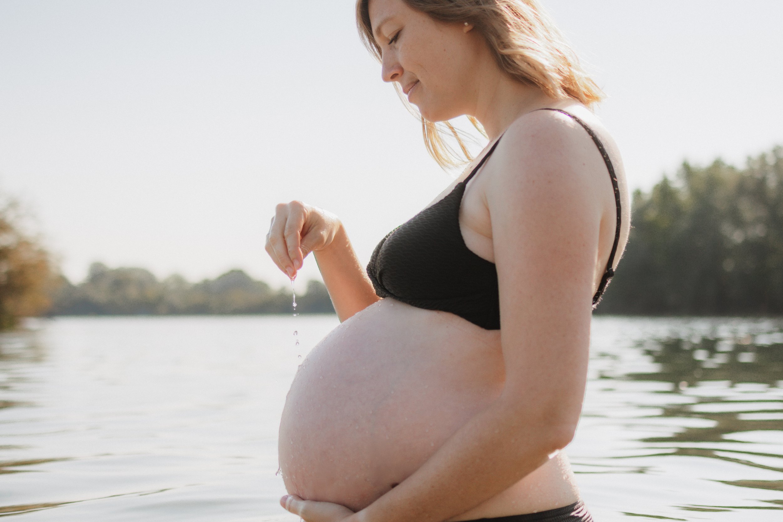 Schwangere Frau im Wasser, die Wasser über ihren Bauch tröpfelt, bei Sonnenuntergang oder -aufgang.