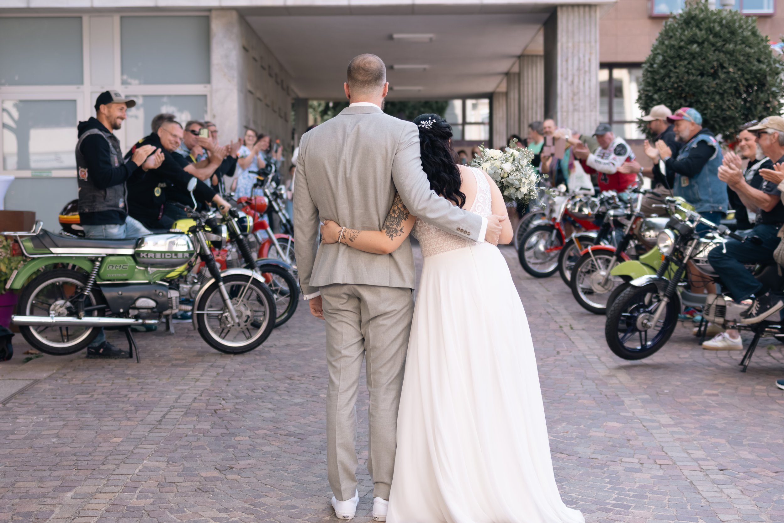 Ein Paar in Hochzeitskleidung steht vor einer Gruppe von Motorradfahrern, die applauding und das Brautpaar bewundern.