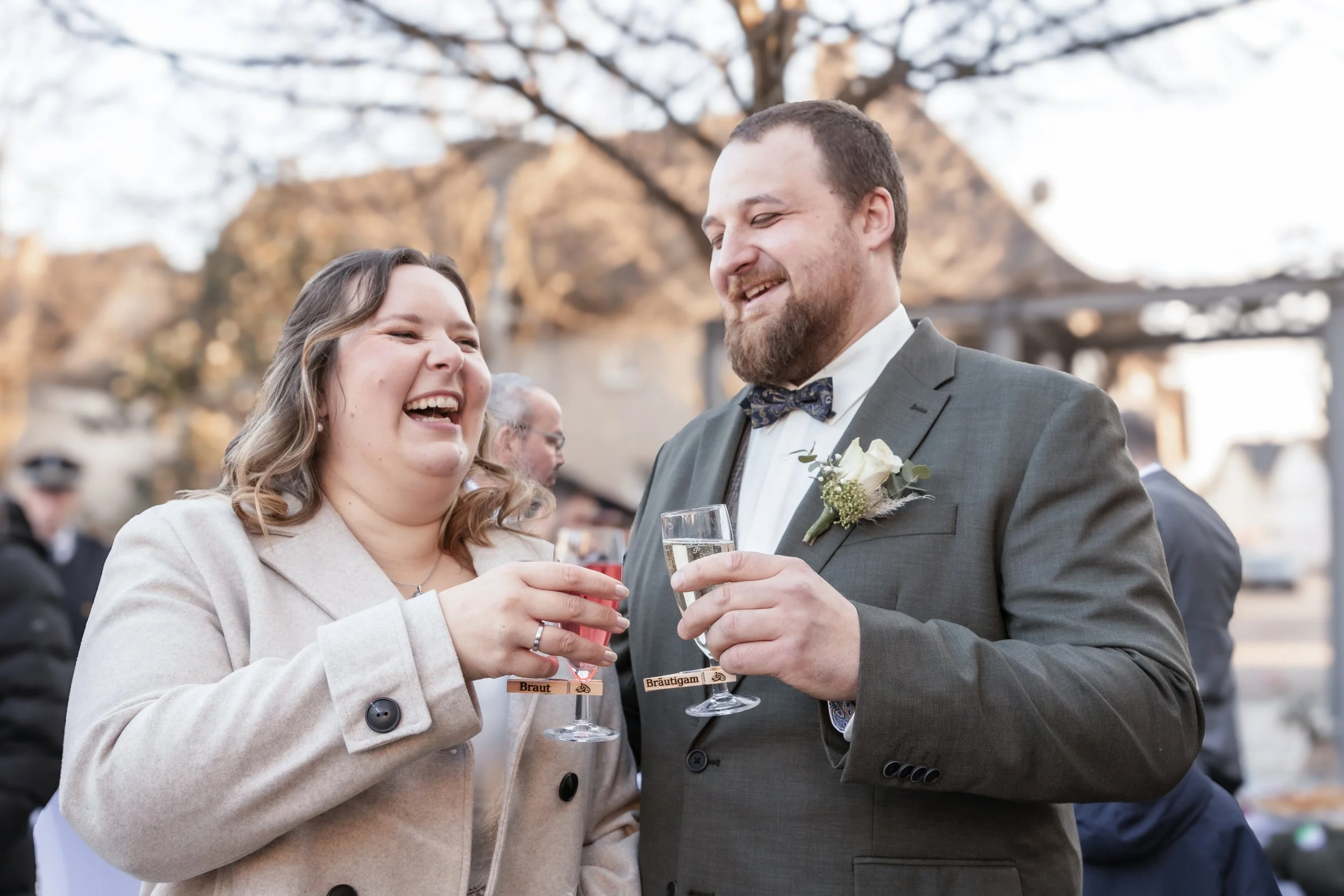 Ein lachendes Paar bei einer Hochzeit, das auf einem Fest mit Gläsern anstößt, im Freien bei Sonnenuntergang, im Hintergrund andere Gäste und Bäume.