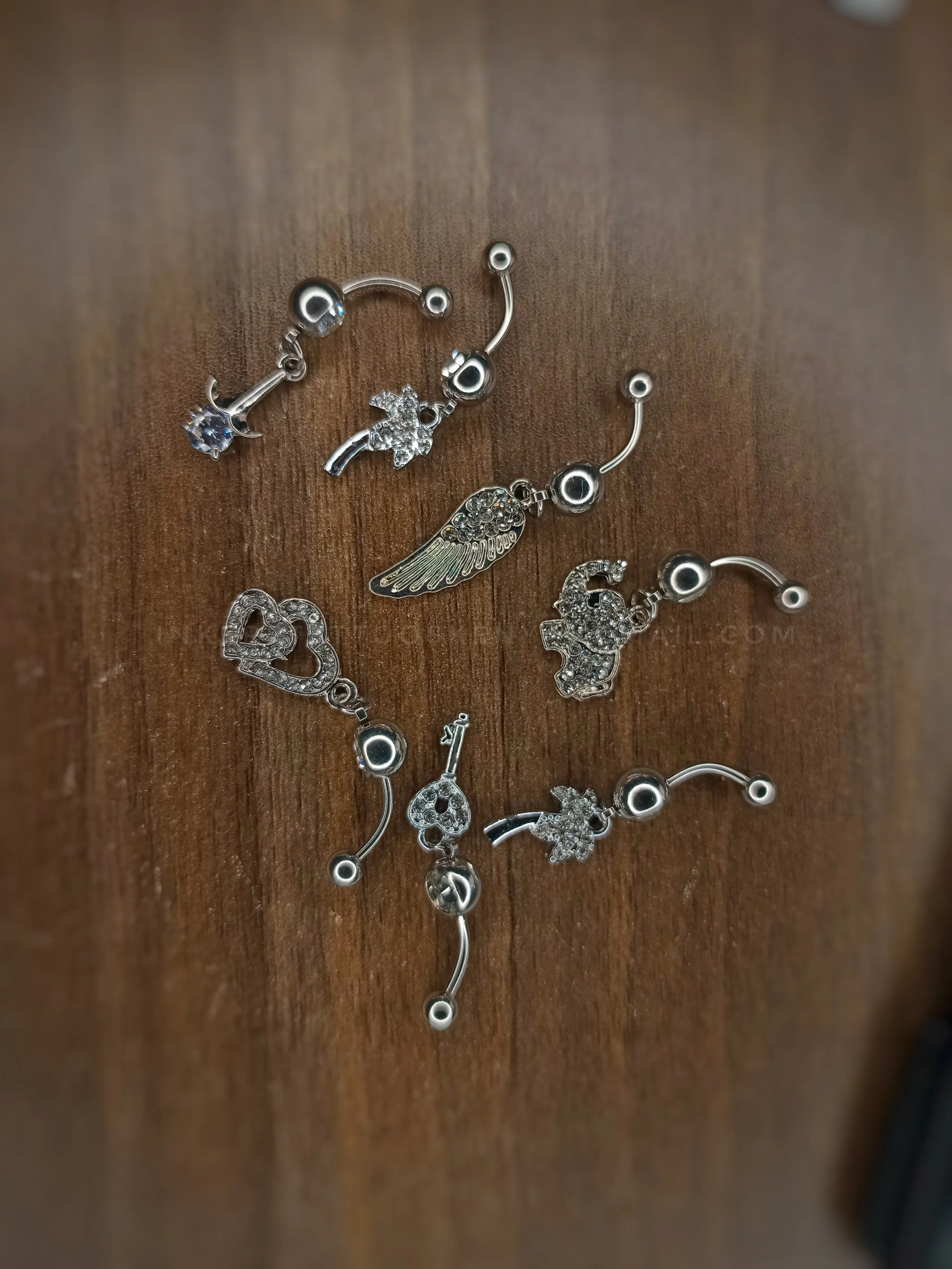 A collection of silver belly button rings with various charms, including a star, a wing, and hearts, laid out on a wooden surface.