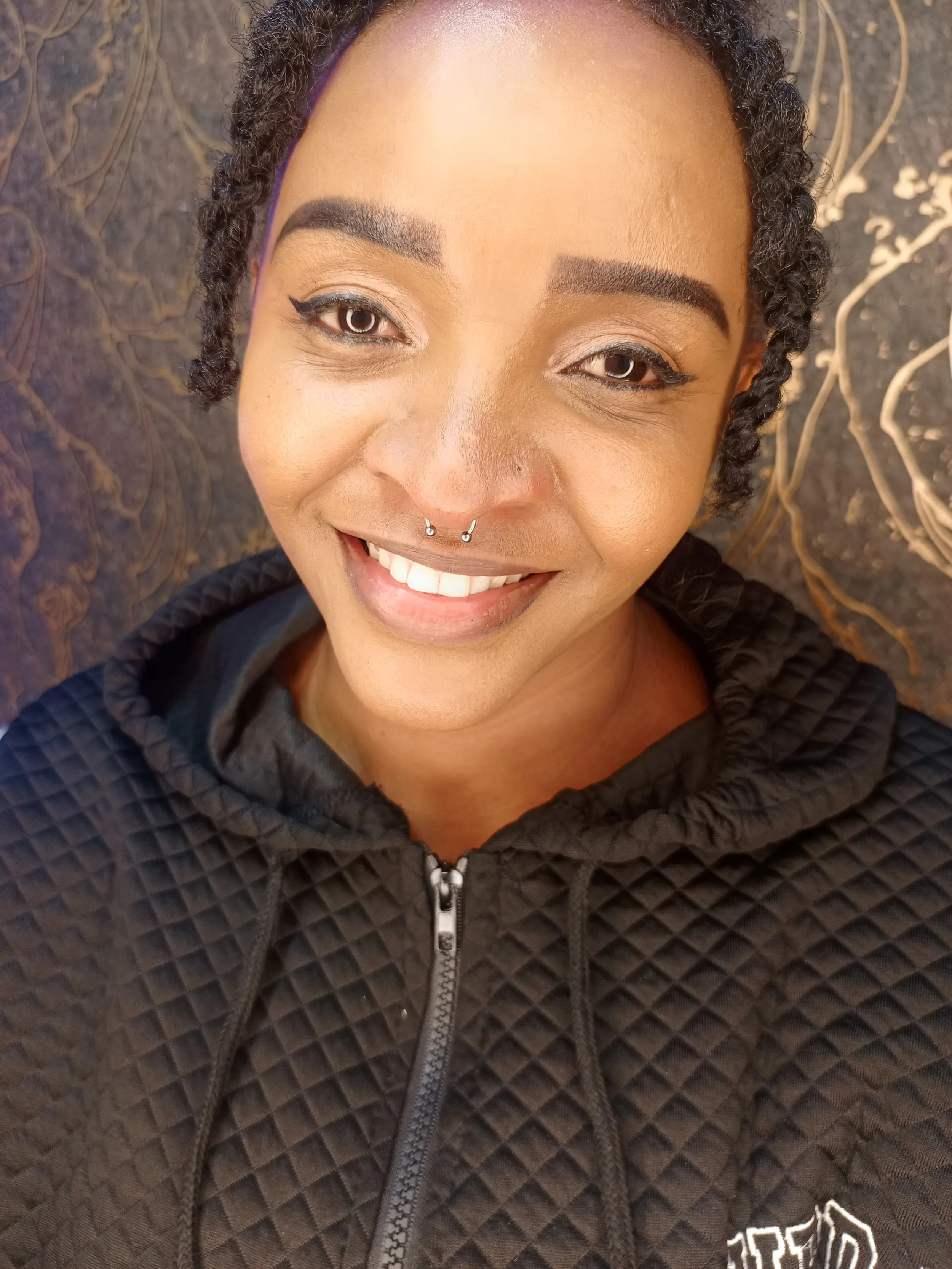A woman with short, curly hair, wearing a black quilted hoodie, smiling at the camera, lying on a dark patterned surface.