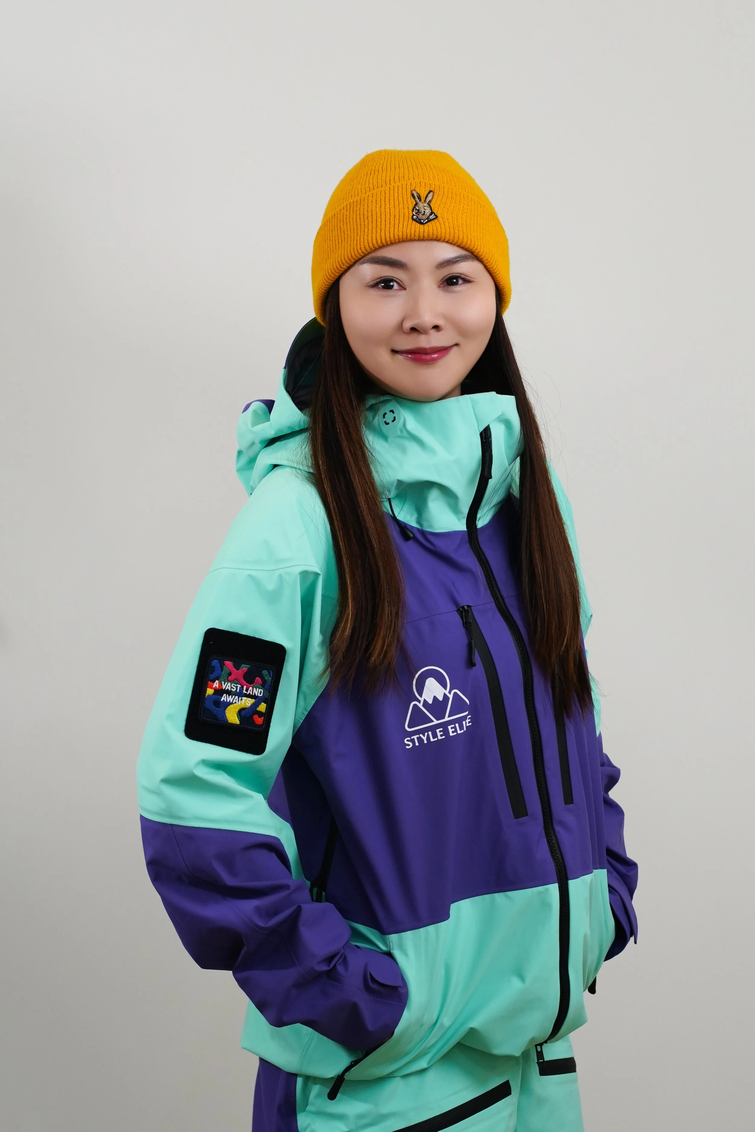 A woman in colorful winter jacket and black beanie standing outdoors on snow-covered mountain with ski lifts in background.