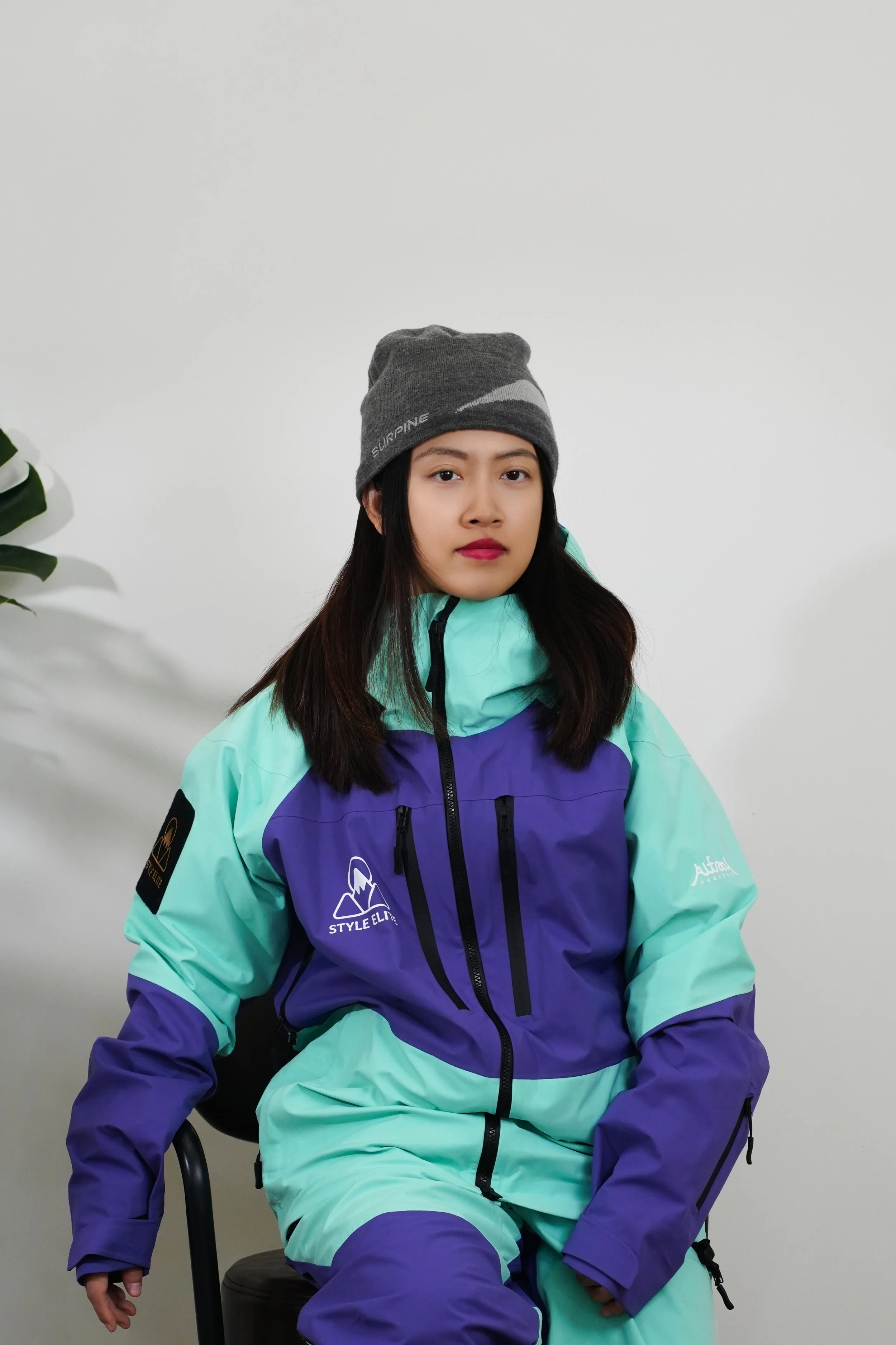 A young woman wearing a beige beanie, a purple and green jacket, standing outdoors with her arms crossed, looking at the camera. The background shows a park with trees and a walkway.