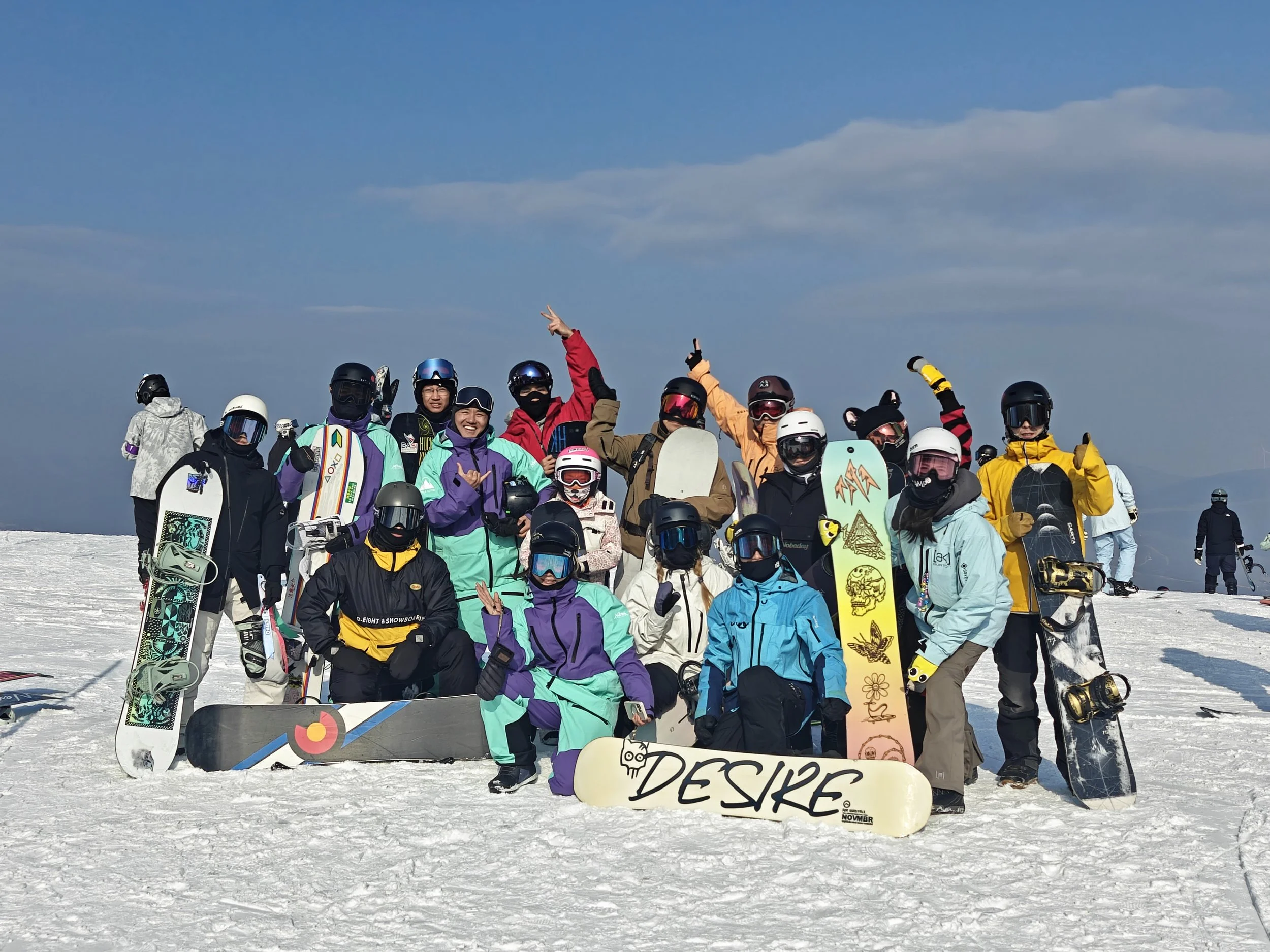 Group of snowboarders and skiers posing together on a snowy mountain with clear blue sky, holding snowboards and skis, some making peace signs and wearing colorful winter gear.