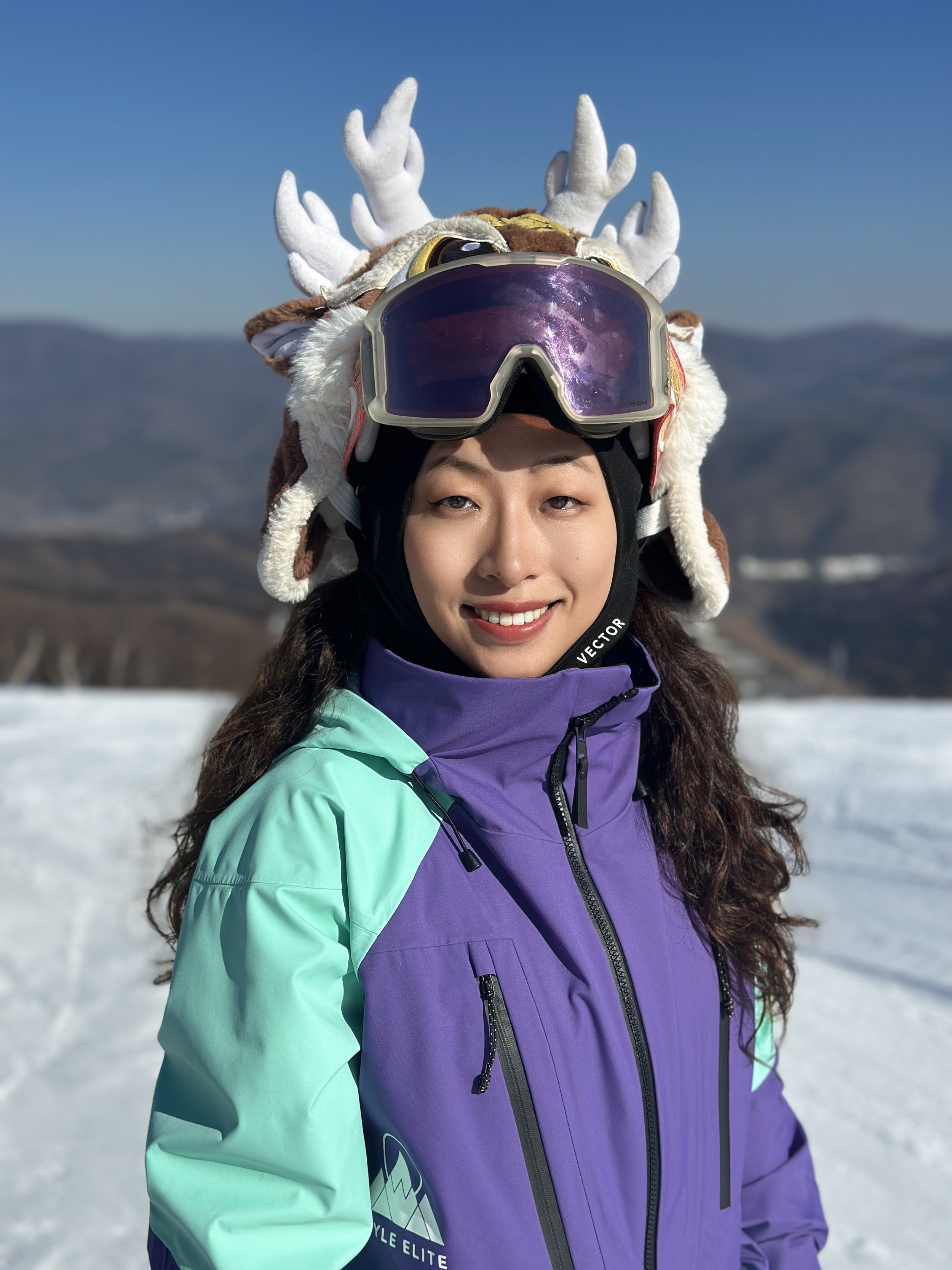 Woman in a purple and teal ski jacket smiling in a snowy mountain landscape, wearing ski goggles and a reindeer hat with antlers.