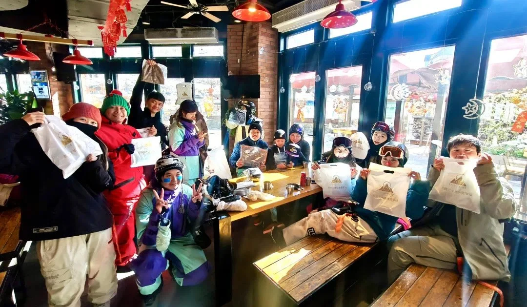 A group of people, mostly children, sitting and standing around a wooden table inside a restaurant or cafe. They are holding white gift bags and wearing winter clothing, including hats, gloves, and jackets. Some are smiling, and one child in front is making a peace sign. The atmosphere is festive with holiday decorations and large windows letting in natural light.