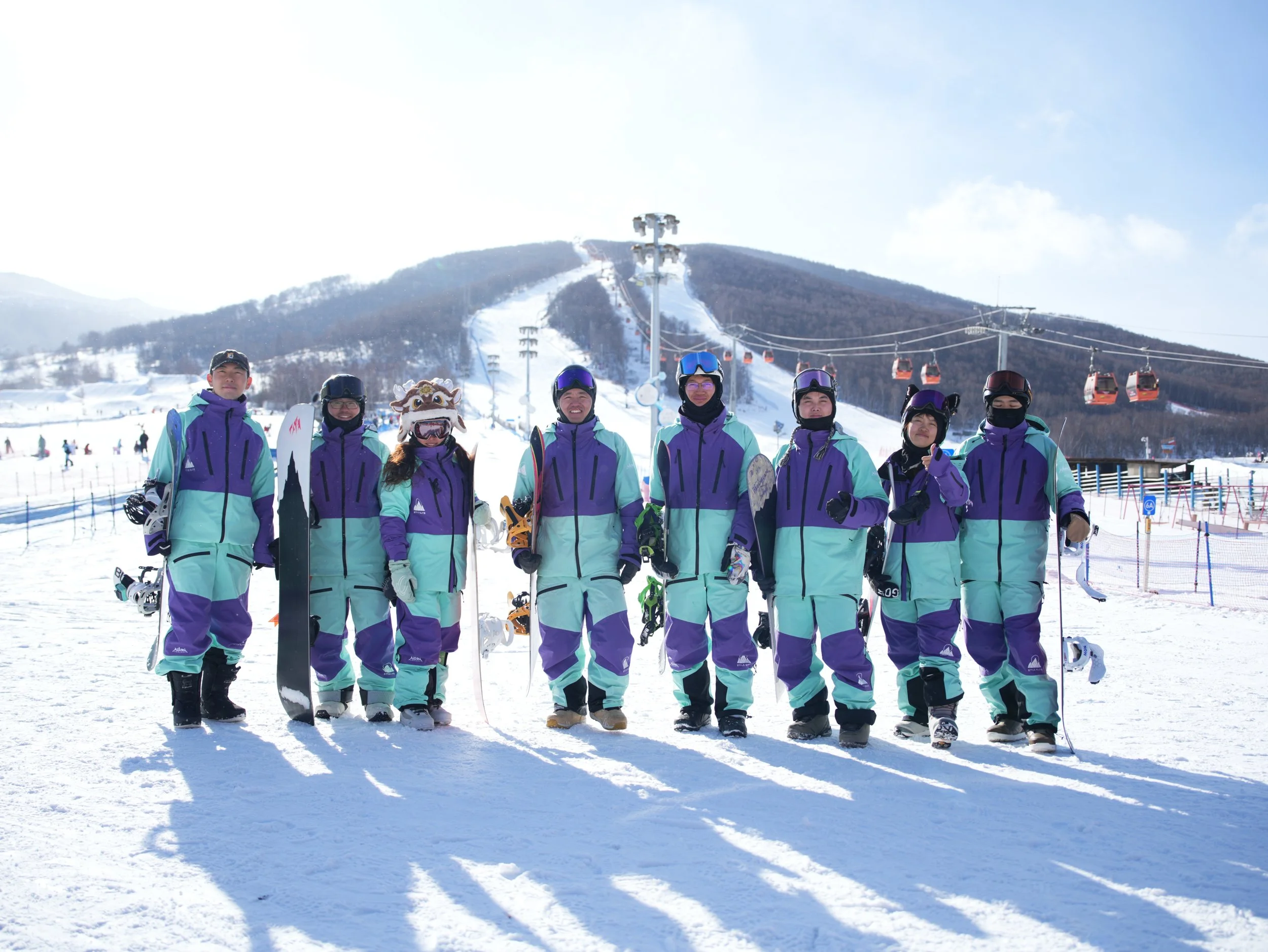 Group of nine people in colorful skiing outfits standing on snow at a ski resort, with ski lifts and slopes in the background.