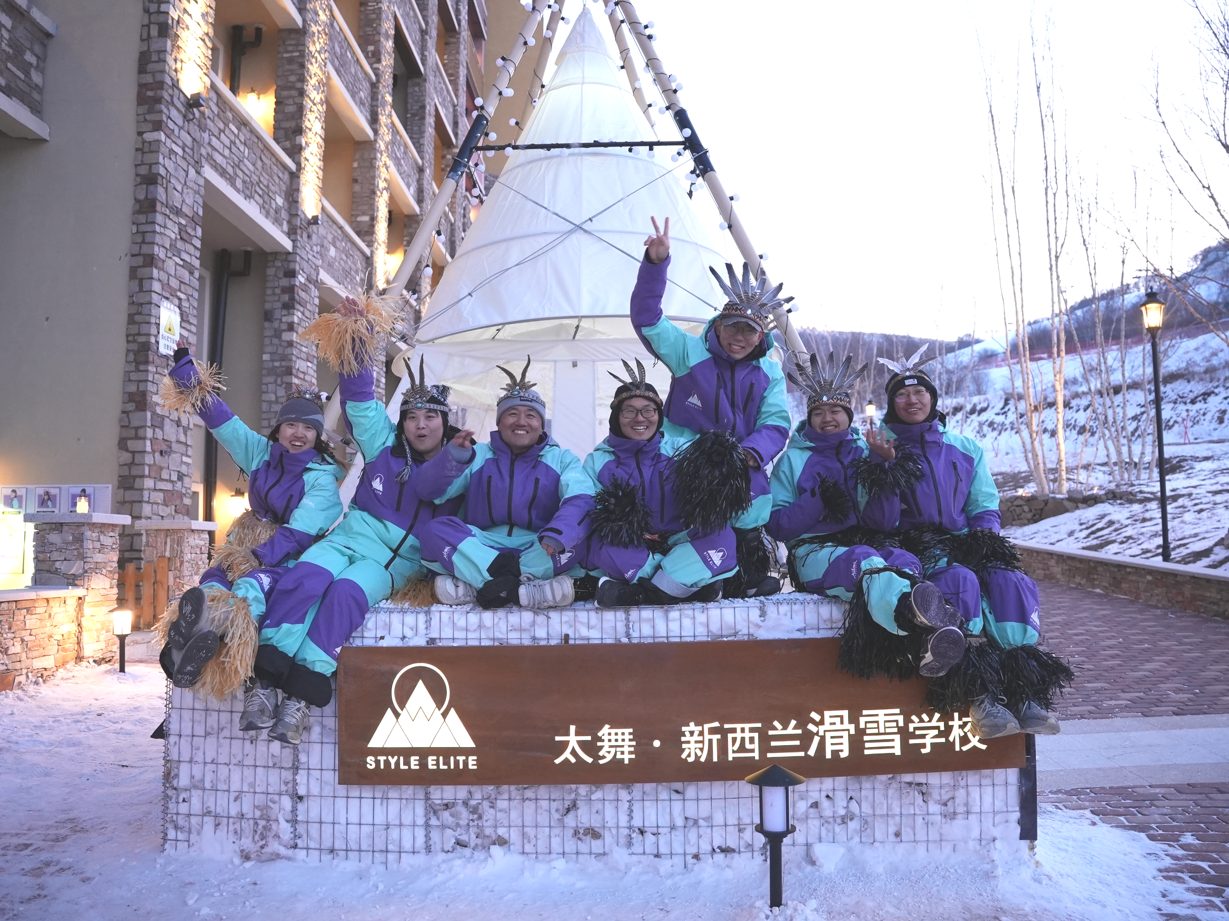 A group of seven people in colorful ski outfits and Native American headdresses sitting on a decorated snow platform outdoors, with a teepee behind them and snow-covered mountains and trees in the background.