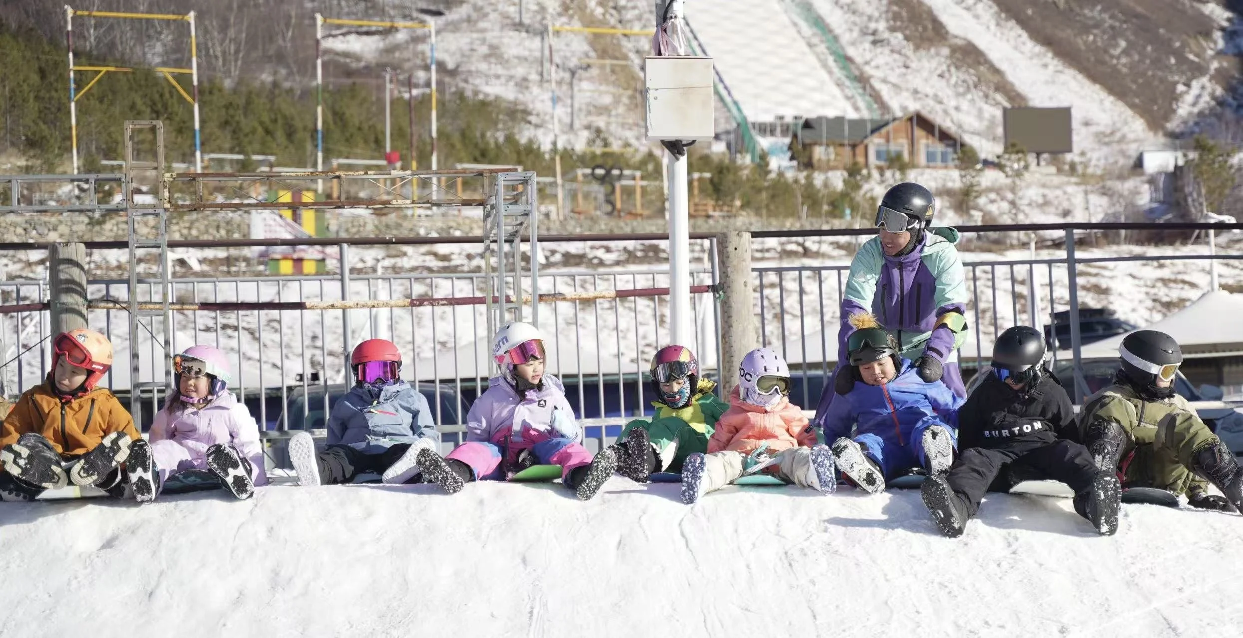 A group of children in colorful winter gear and helmets sitting on the snow, with an instructor standing next to them, outdoors in a snowy mountain area.