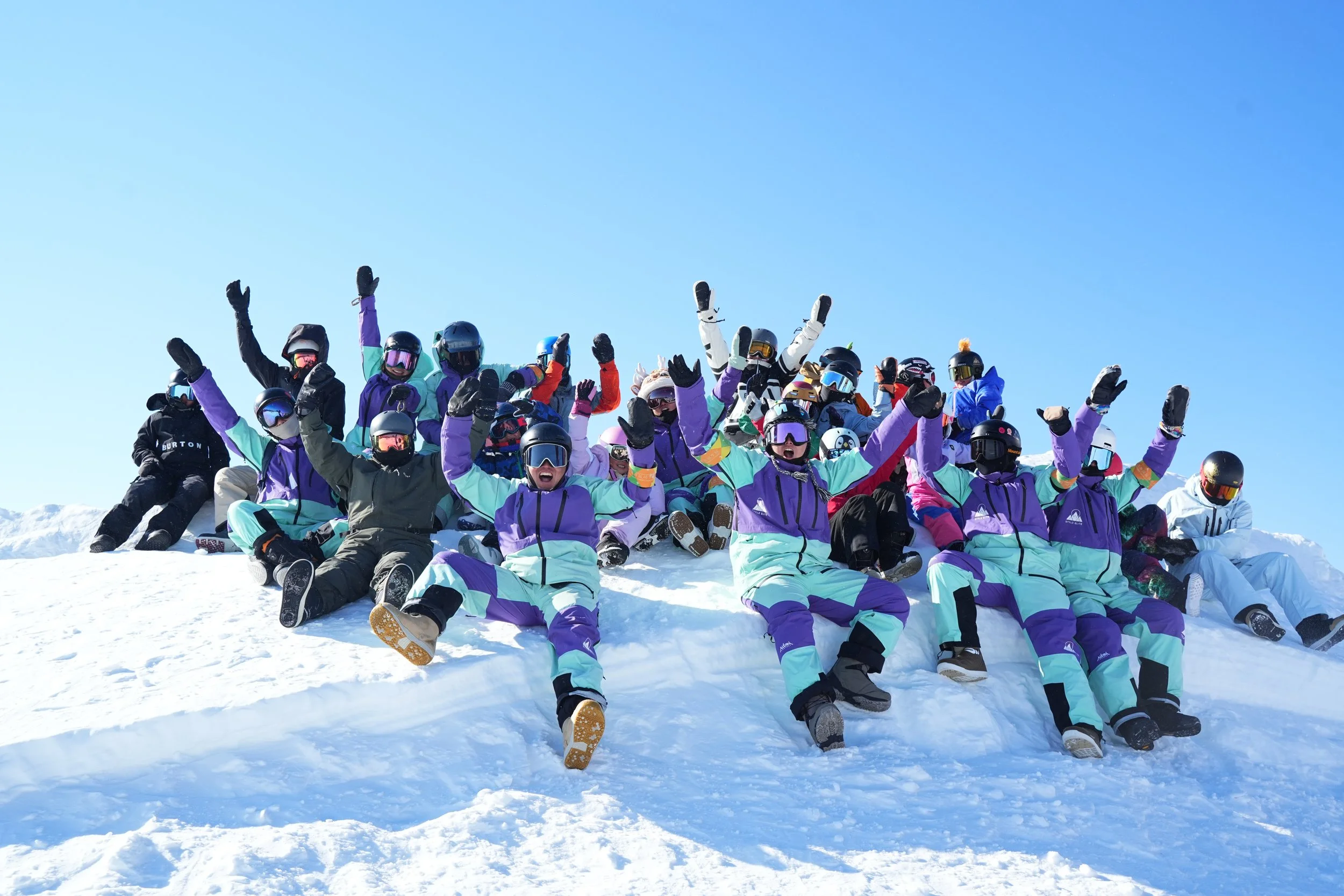 A large group of people wearing winter clothing and ski helmets, sitting and raising their arms on a snowy mountain slope under a clear blue sky.