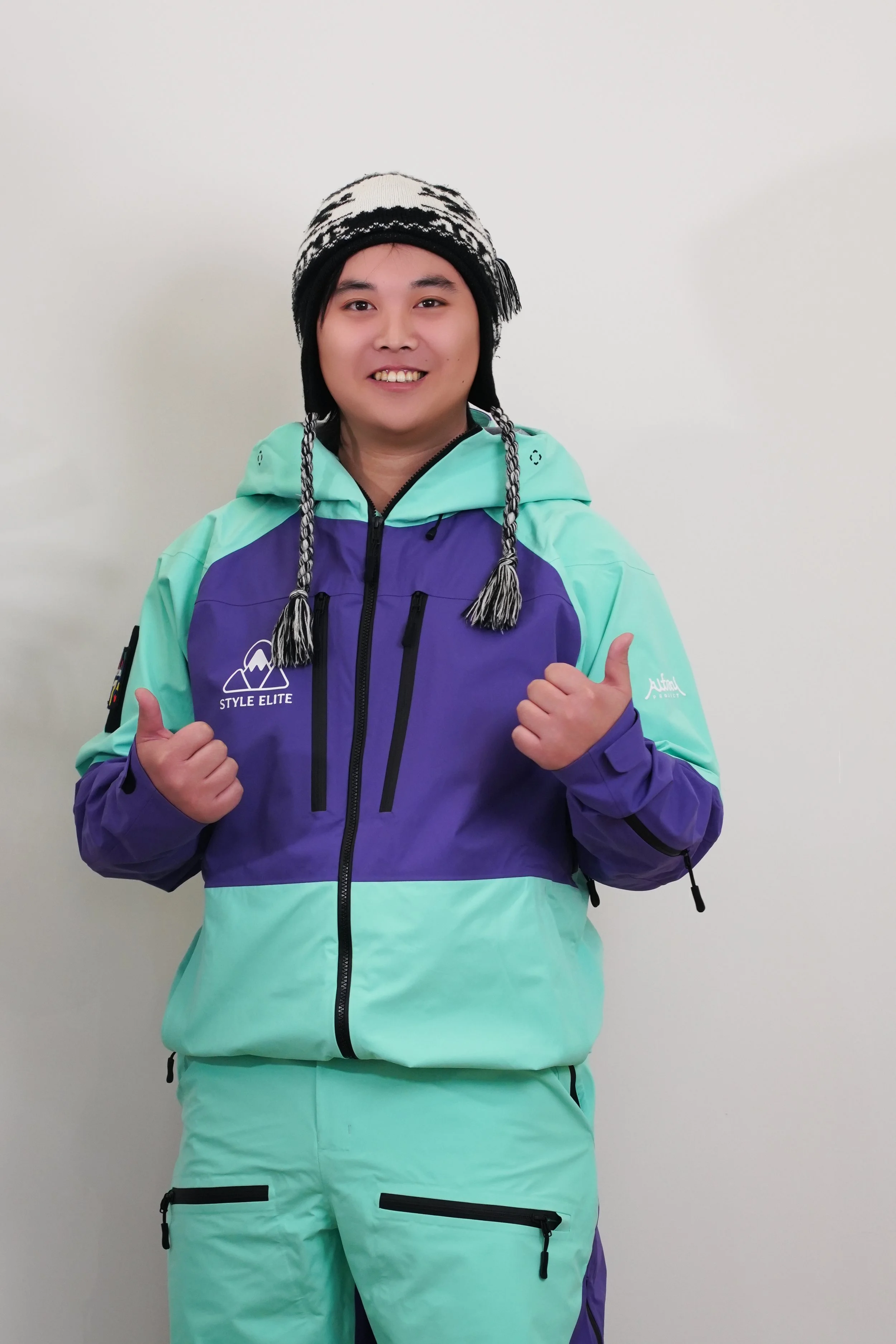 A person wearing a ski helmet and goggles, smiling and giving thumbs up, standing indoors with decorative skateboards and pink flowers in the background.