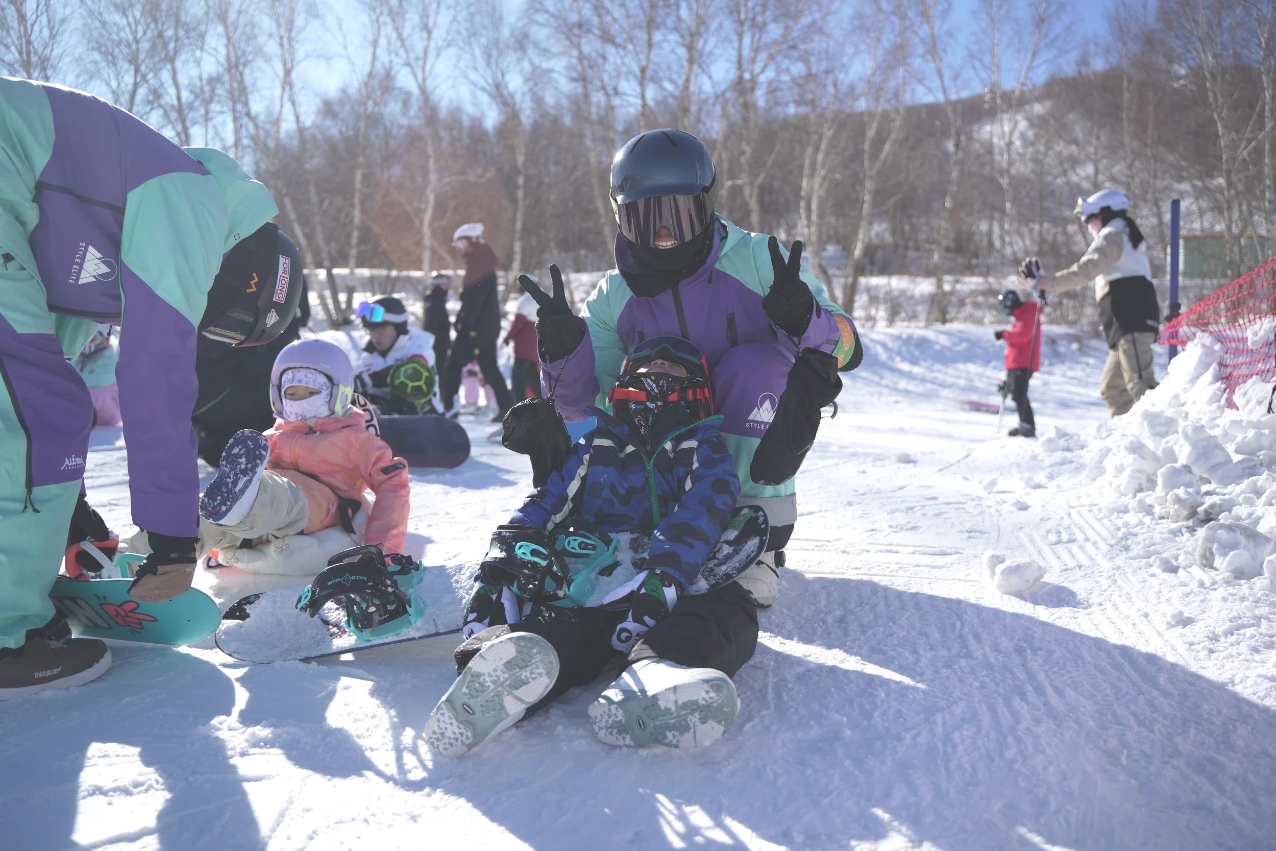 A group of people, including children and adults, at a snowy ski resort. Some are sitting and preparing their ski equipment, while others are standing or skiing in the background. The person in the center is sitting on the snow with a child, both dressed in ski gear, and waving at the camera.