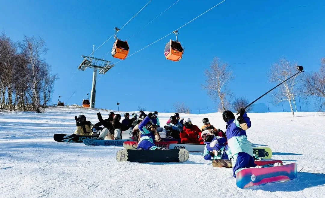 Group of snowboarders and skiers sitting and lying on the snow at the bottom of a ski slope with ski lifts in the background and clear blue sky.