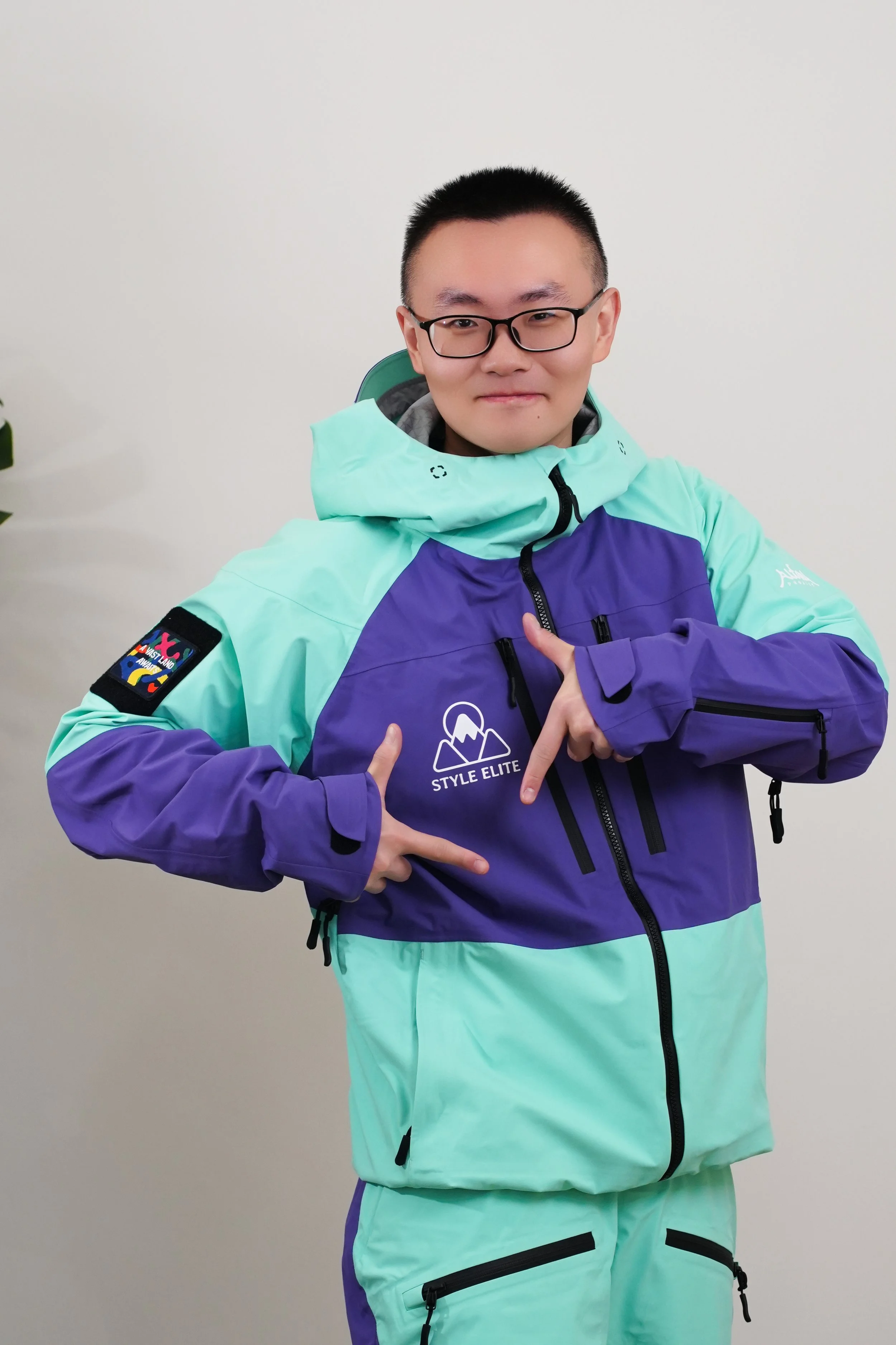 A man wearing a black ski helmet, black glasses, and a colorful ski jacket with purple and mint green colors, standing on snow with a blurred snowy ski resort background.