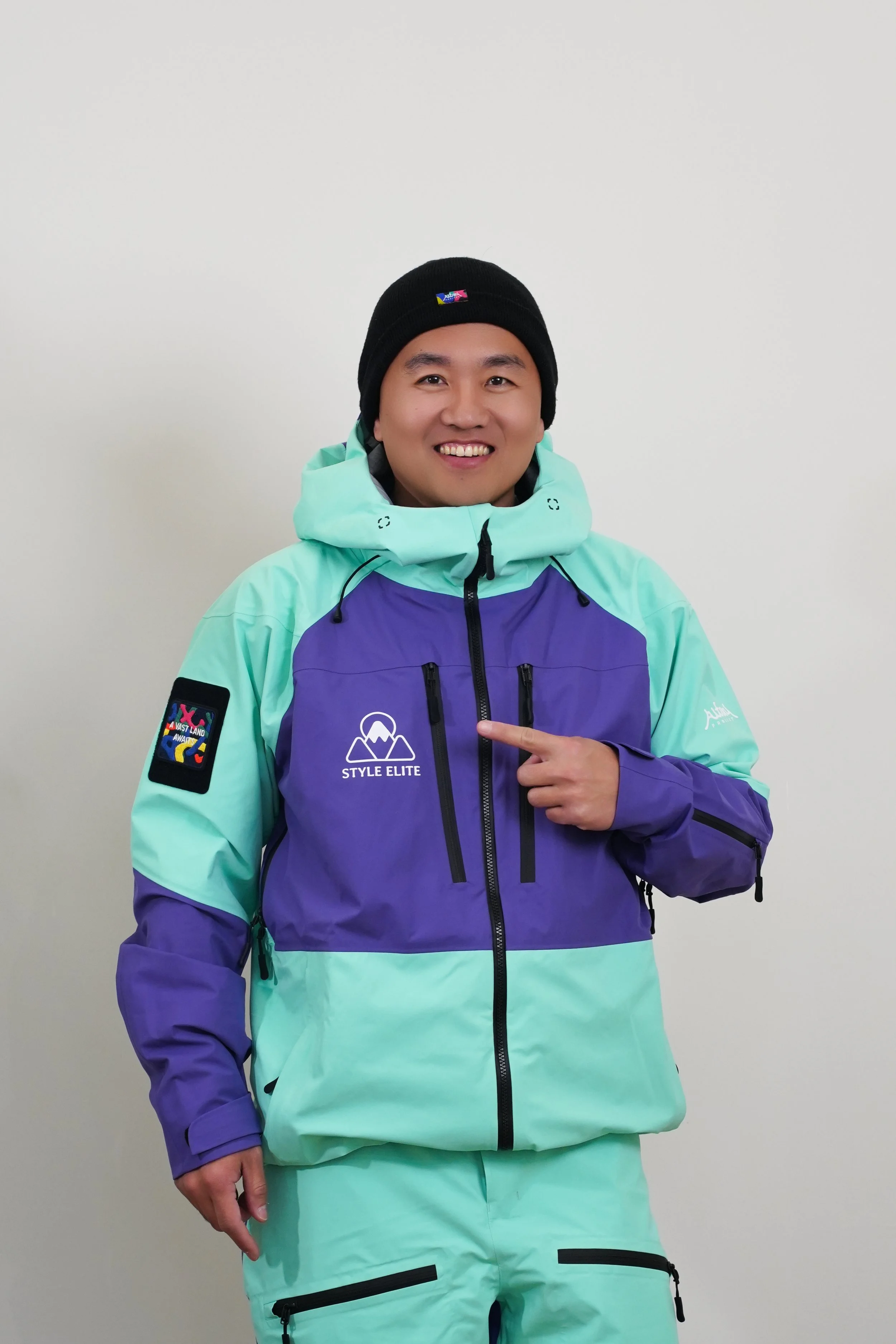 A man wearing a ski helmet, goggles, and a colorful ski jacket standing on snow with a mountain backdrop.