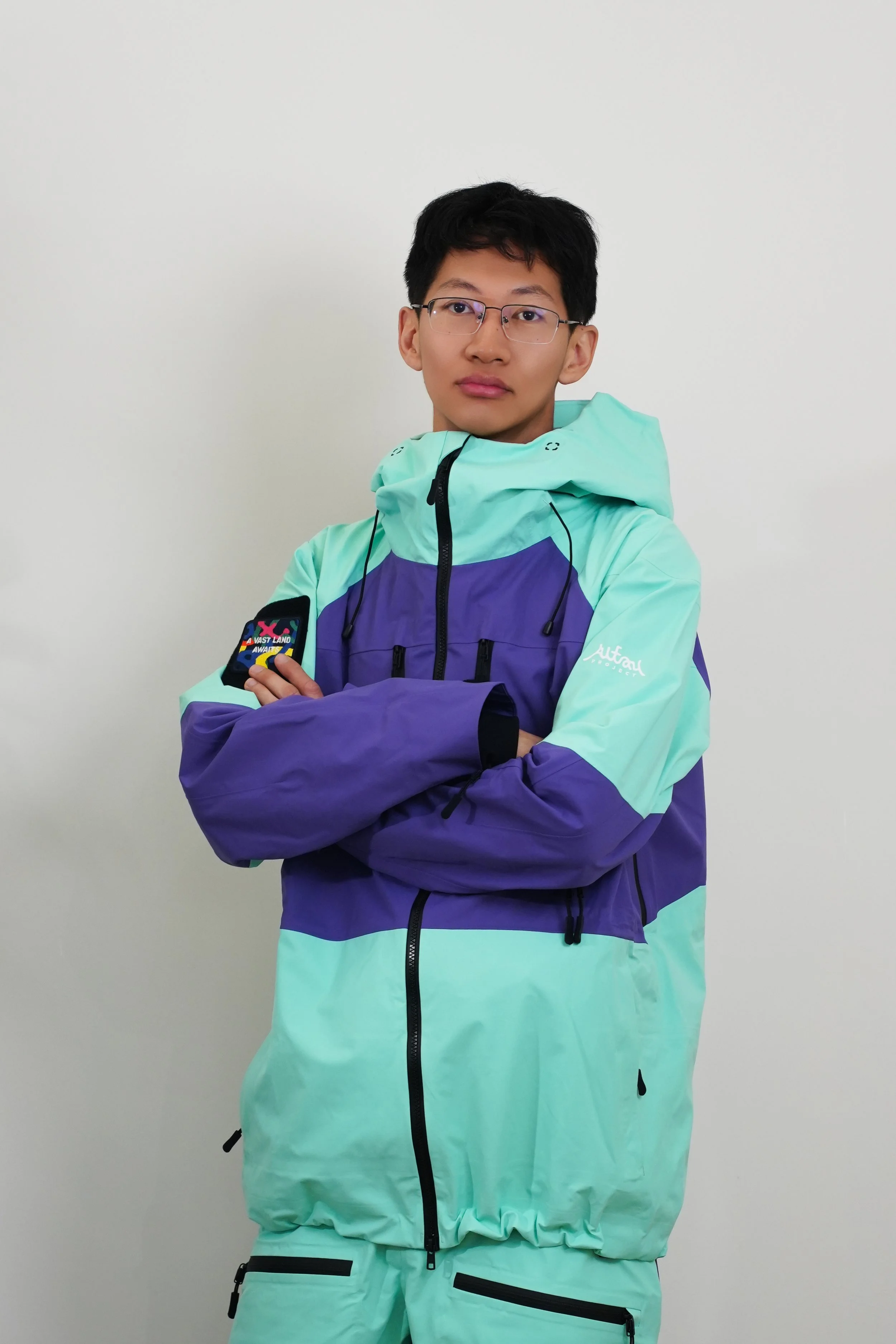 A man wearing ski goggles, a purple and teal ski jacket with a mountain logo, standing on snowy terrain under a clear blue sky.