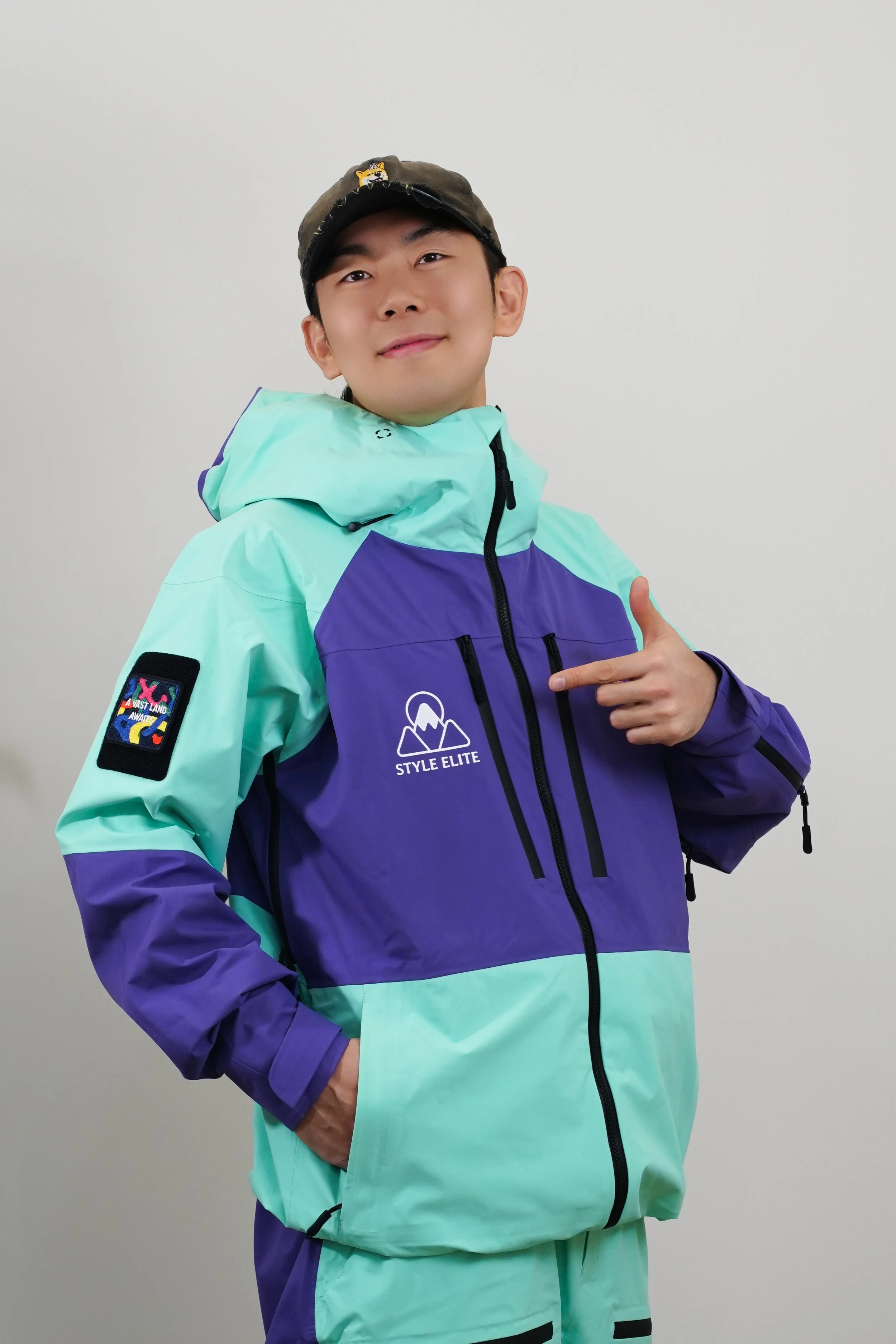 A person in a colorful ski jacket and helmet, standing on snow with a ski lift in the background, sticking out their tongue and smiling.